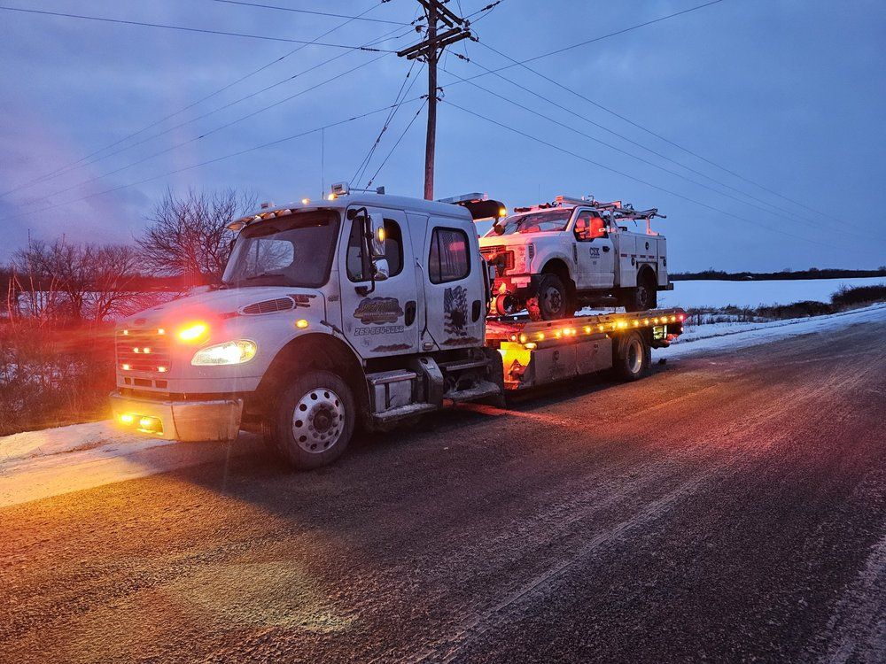 Tow truck with emergency lights carries a utility truck on a snowy road at dusk.