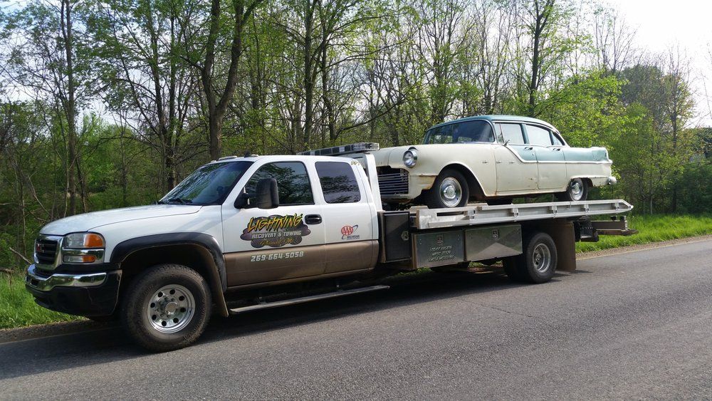 Tow truck carrying a classic two-tone car on a flatbed, parked on a road beside trees.