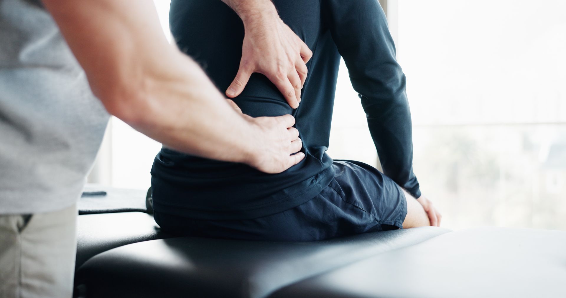 Person holding lower back while seated on a treatment table as a therapist examines their spine