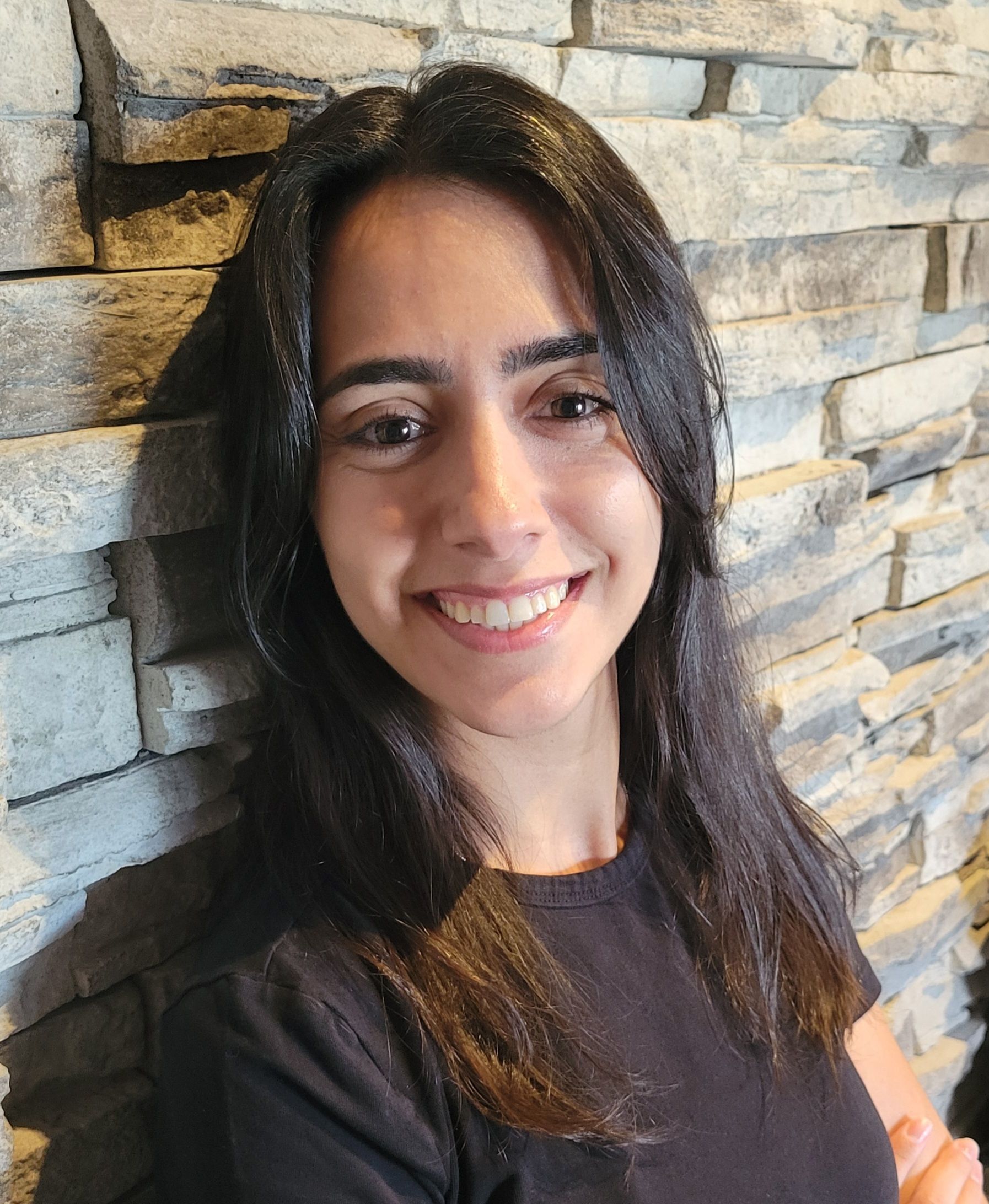 Woman with dark hair smiles at the camera, leaning against a stone wall.