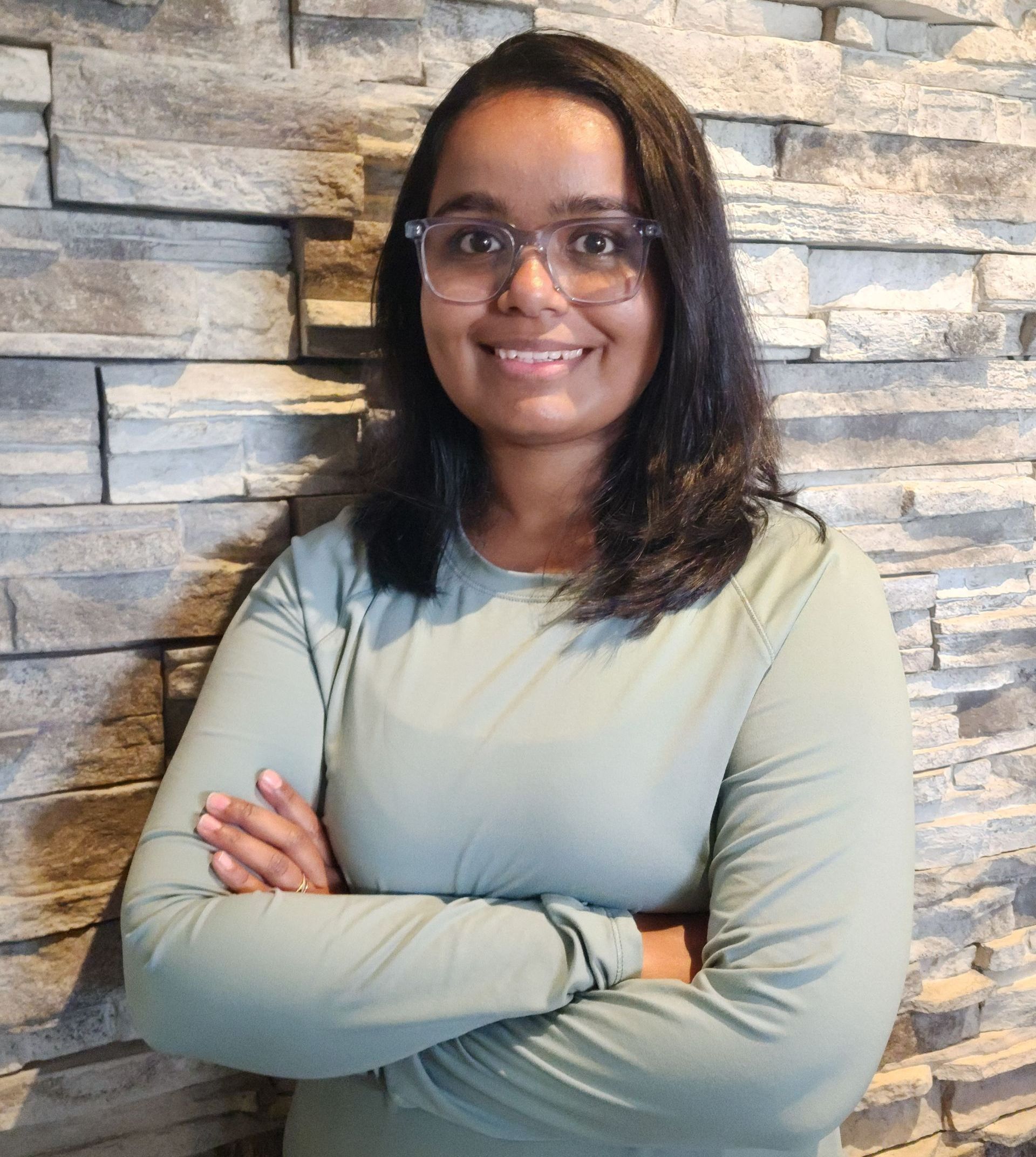 A woman wearing glasses is standing in front of a brick wall with her arms crossed.