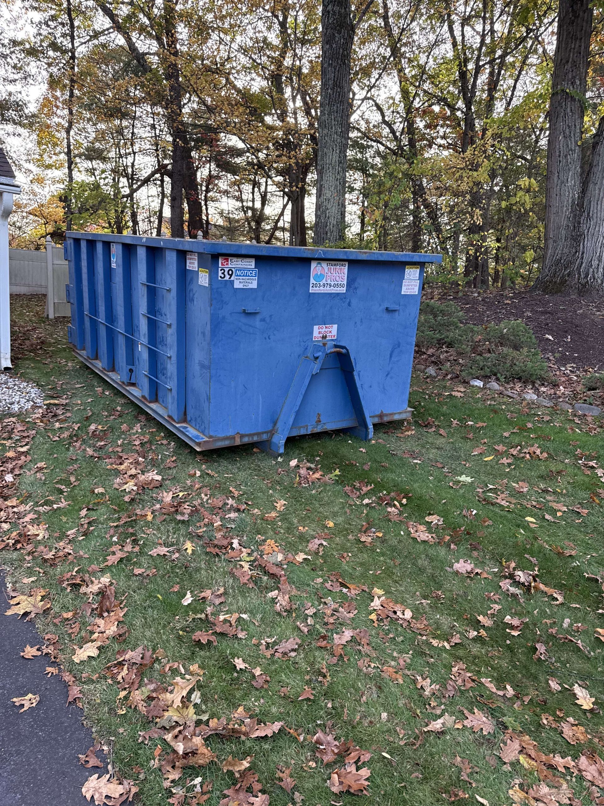 Blue dumpster on green grass, next to a white fence and trees with fall foliage in shelton.