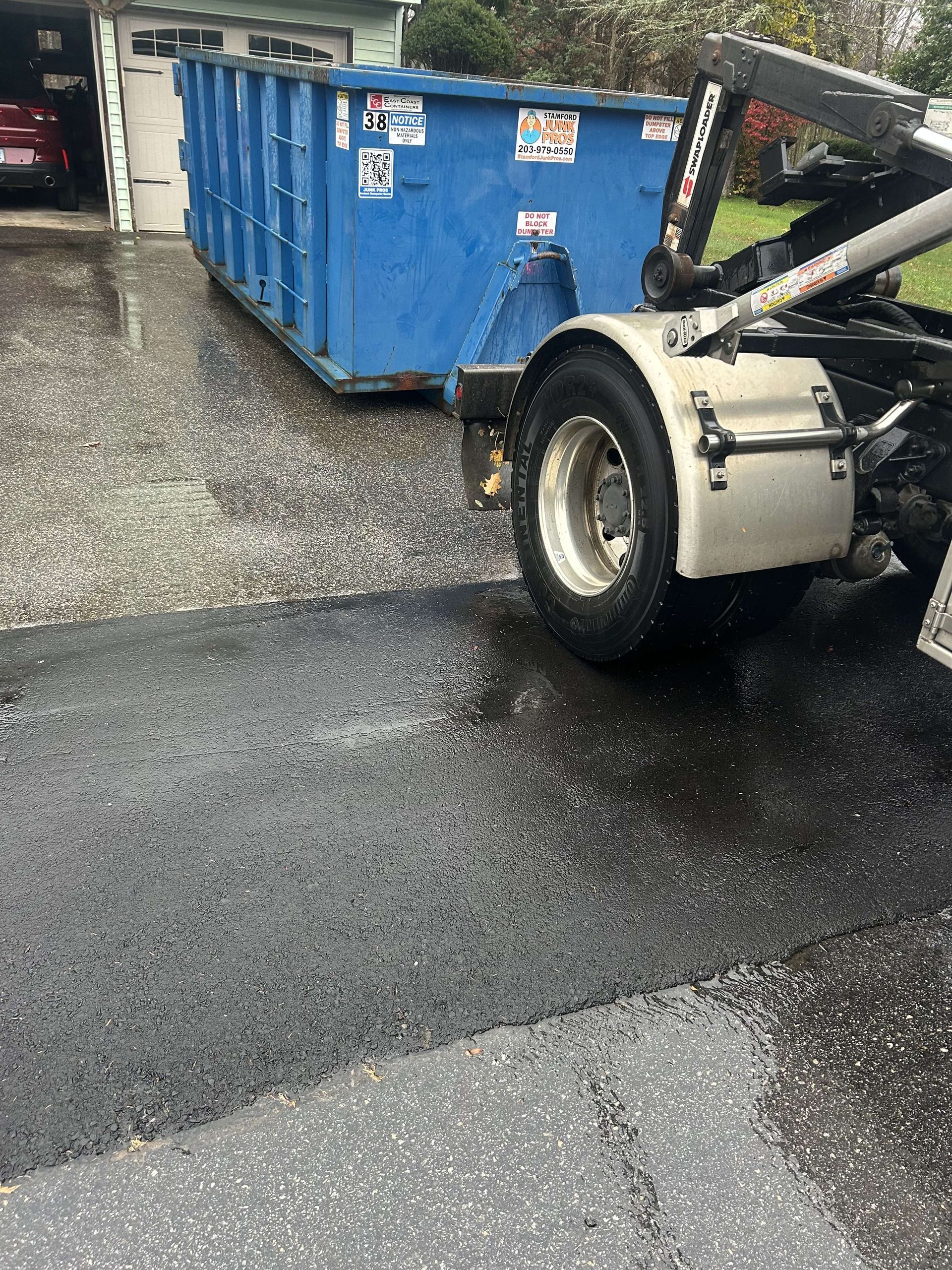 Blue dumpster being hauled by a truck on a wet asphalt driveway, near a garage in Torrington.