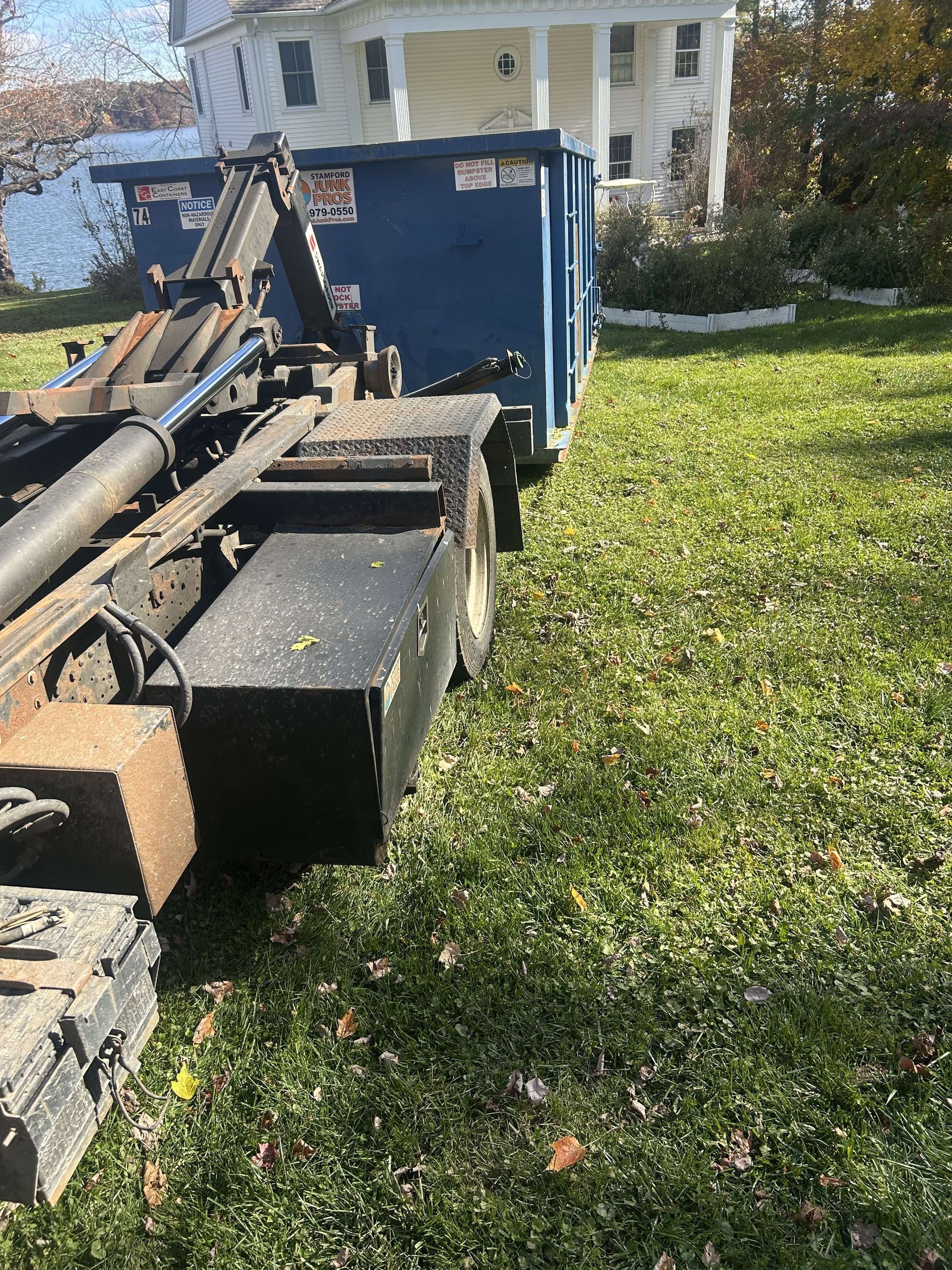 Blue dumpster on a truck, parked on grass in front of a white house.