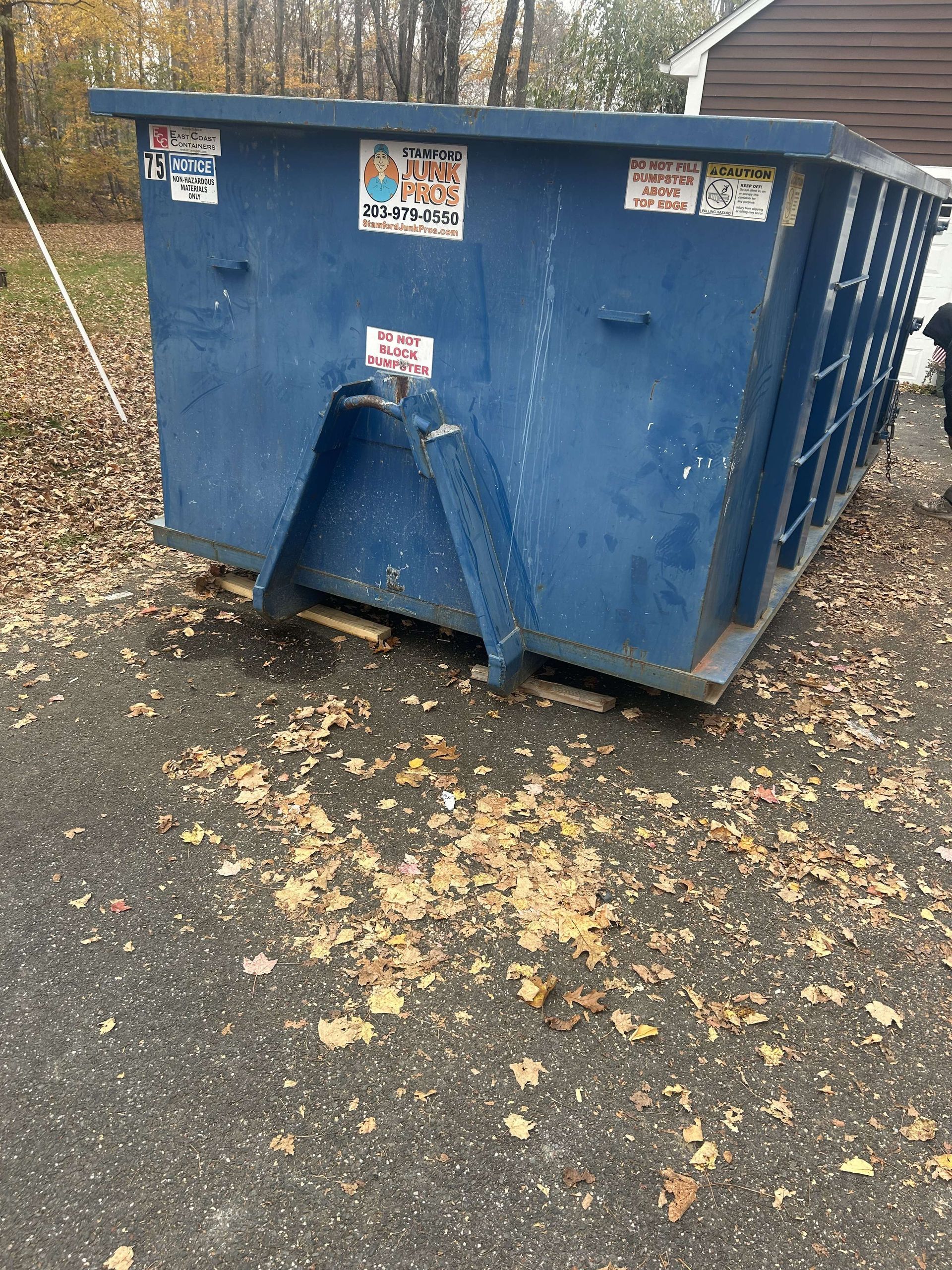 Blue dumpster on a pallet, covered in stickers, outdoors on a paved area with fallen leaves.