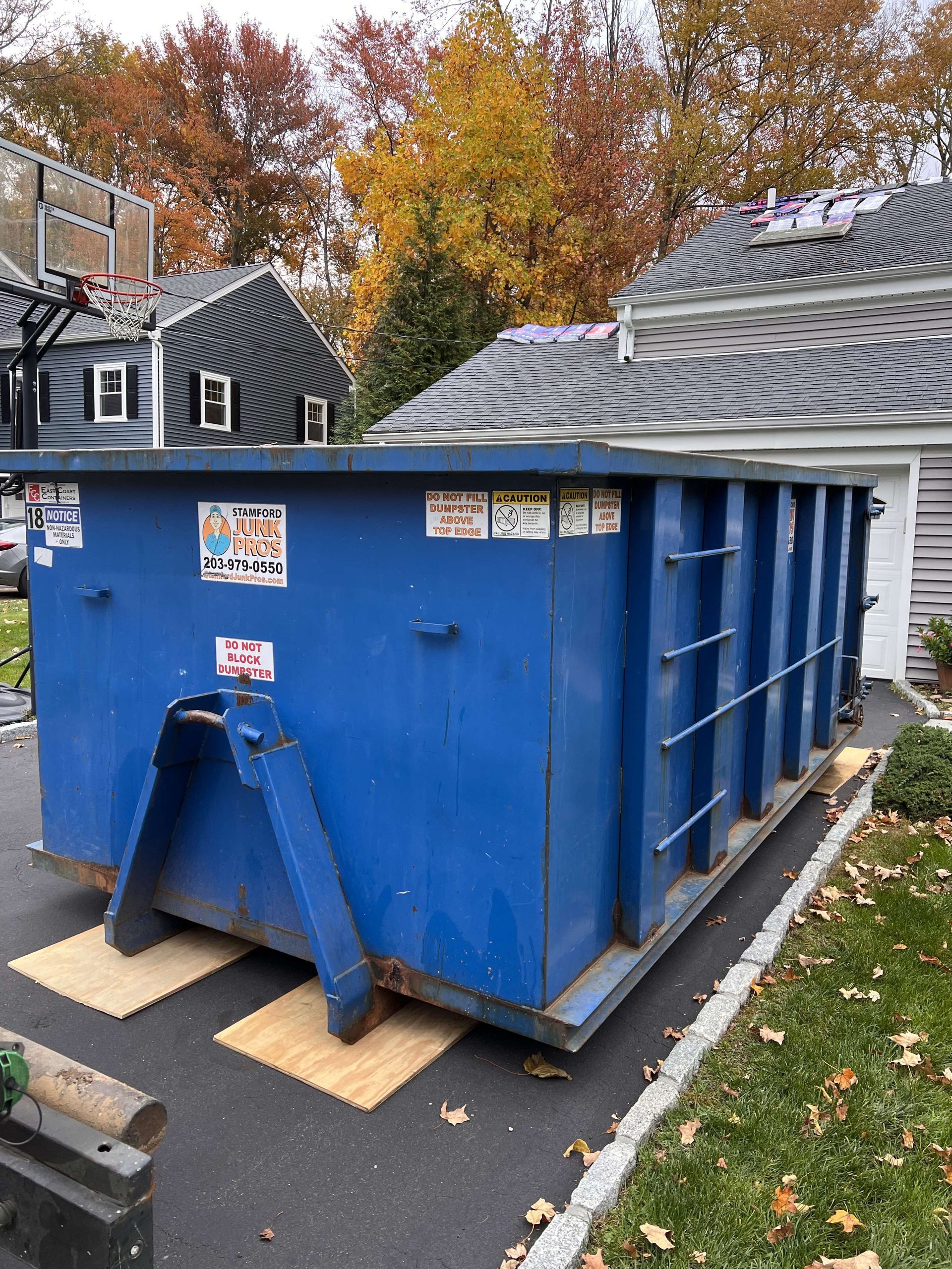 Blue dumpster on a driveway, near a house with roofing work in progress; fall foliage in background.