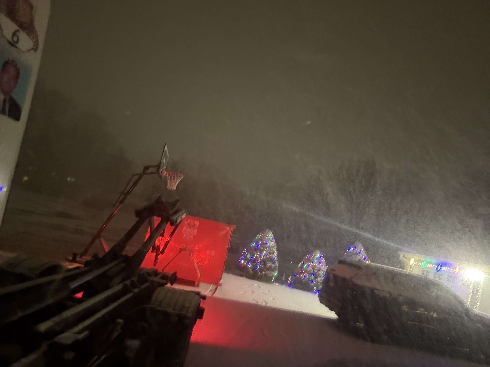Snow-covered street at night with snow falling. A snowblower is visible. Christmas trees are lit up.
