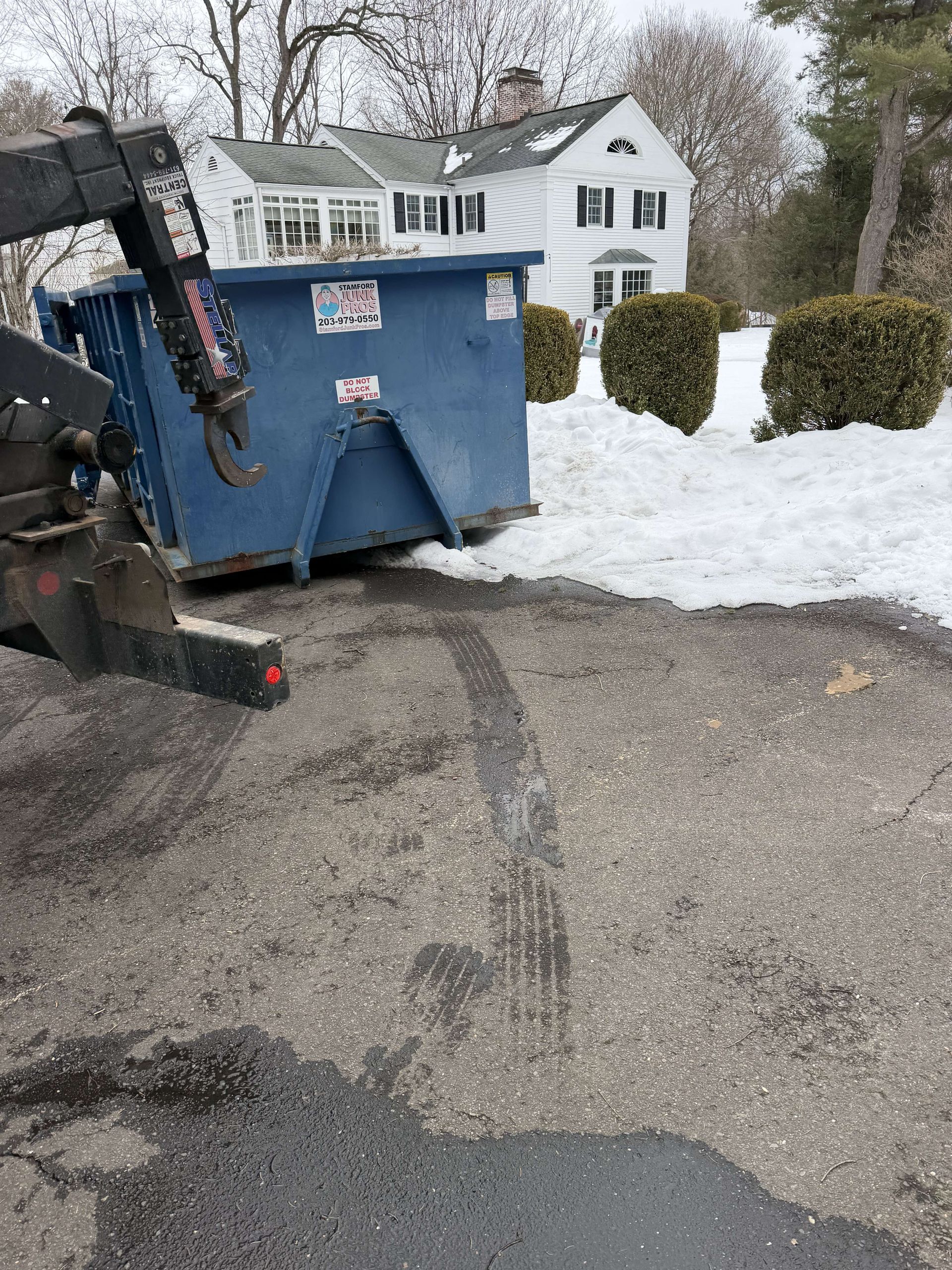 Snow-covered driveway with a blue dumpster and a front loader near a white house and trimmed bushes