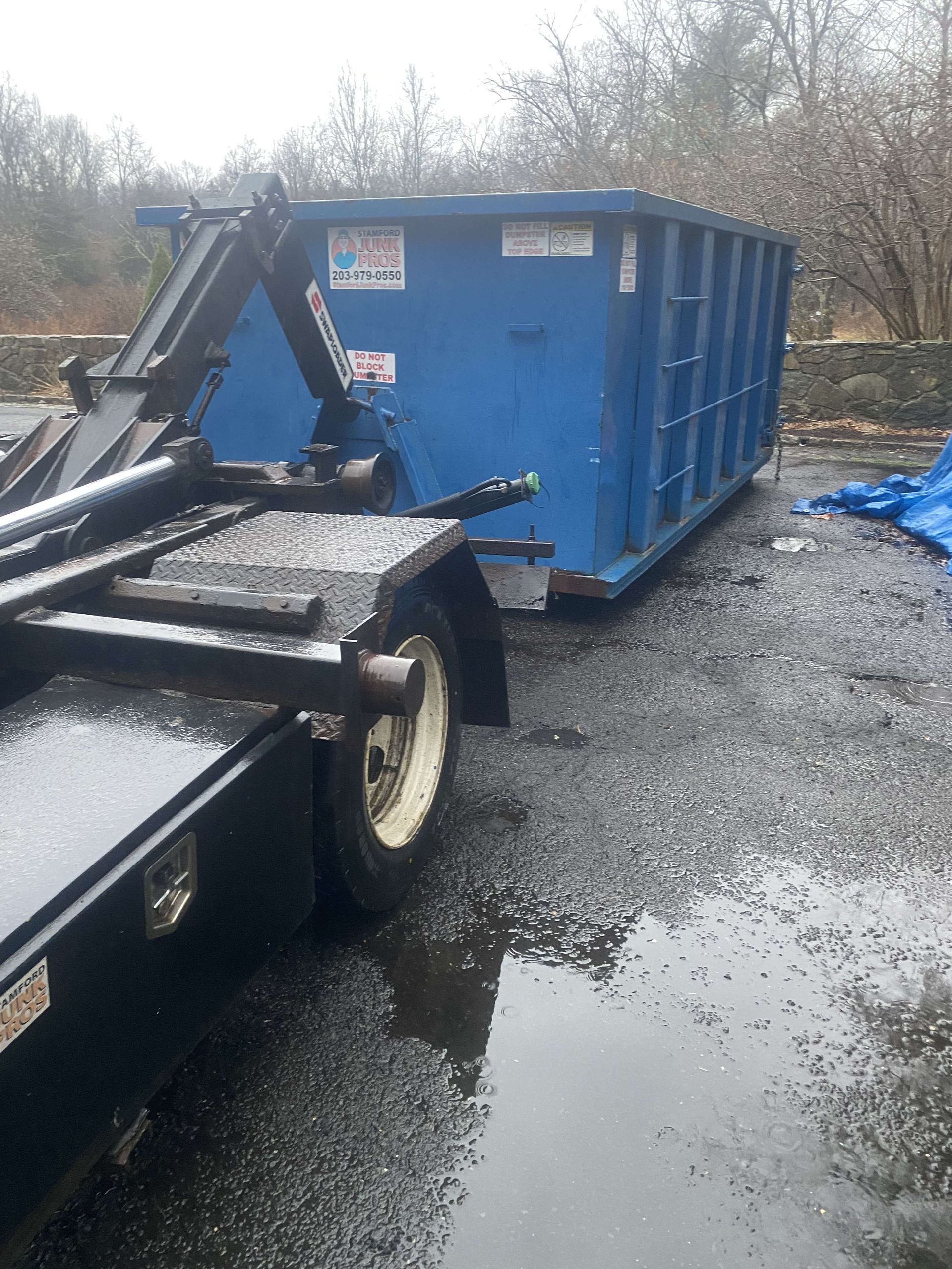 A blue dumpster being loaded onto a trailer on a wet, gray day in milford.