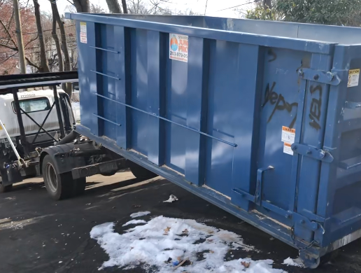 Blue dumpster being lifted by a truck on a snow-covered road.