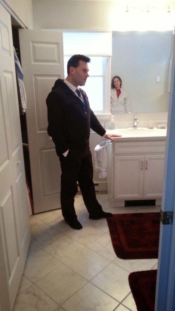 Man in suit stands in bathroom, looking right, hand on vanity. Woman reflected in mirror.