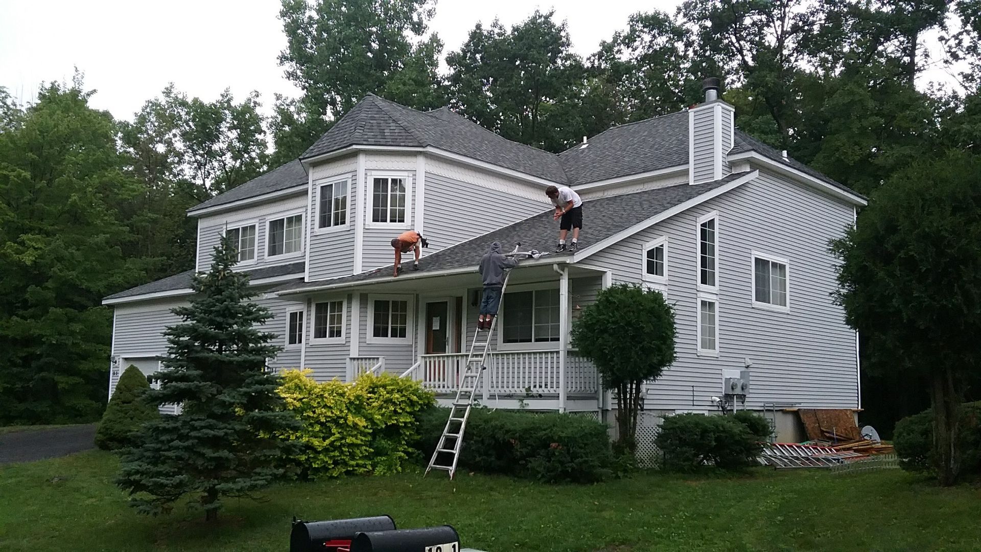 House with two workers on roof, grey siding, trees, ladder.
