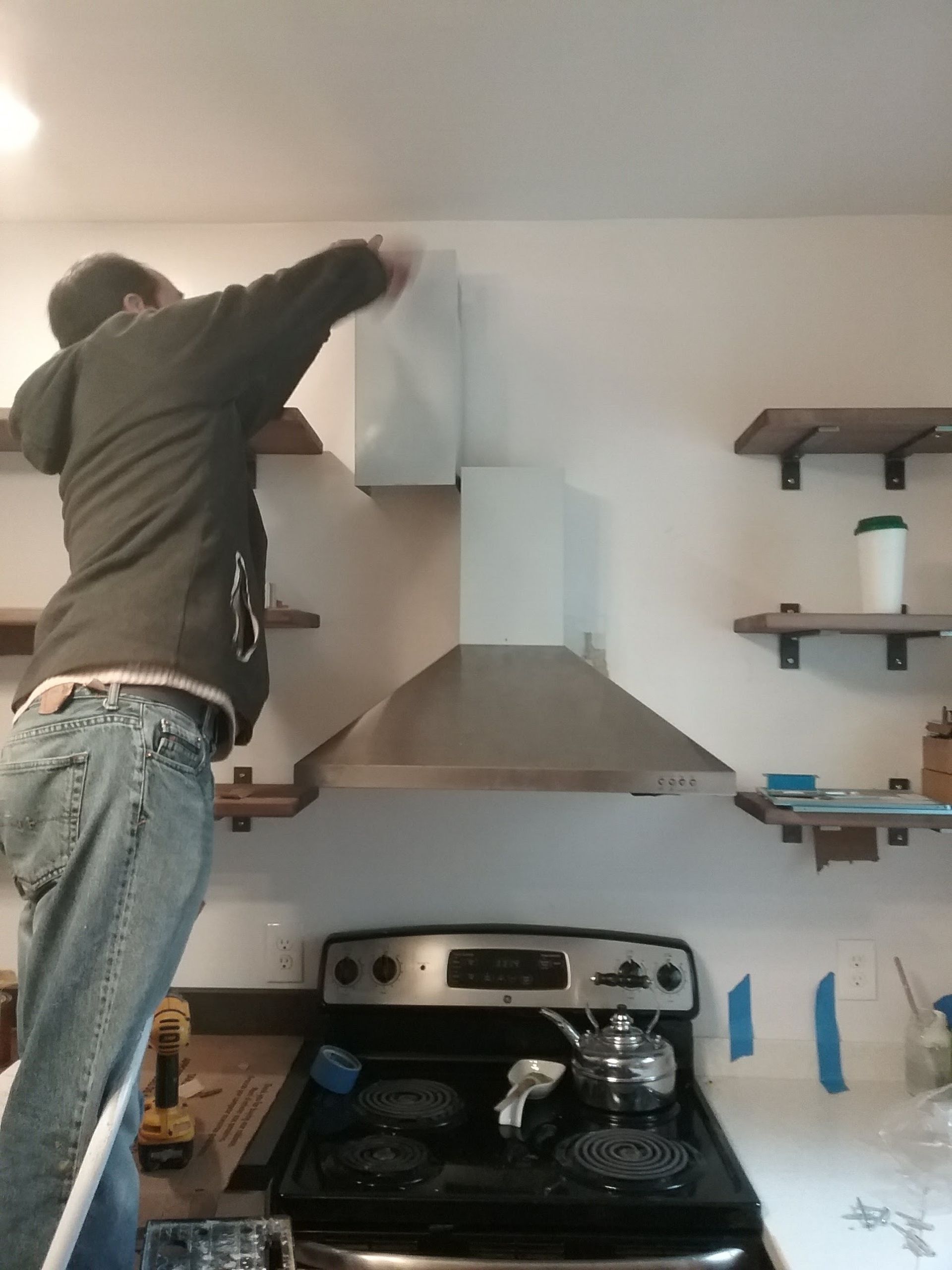 Person on a ladder installing a kitchen vent hood. White wall, stainless steel appliances, open shelves.