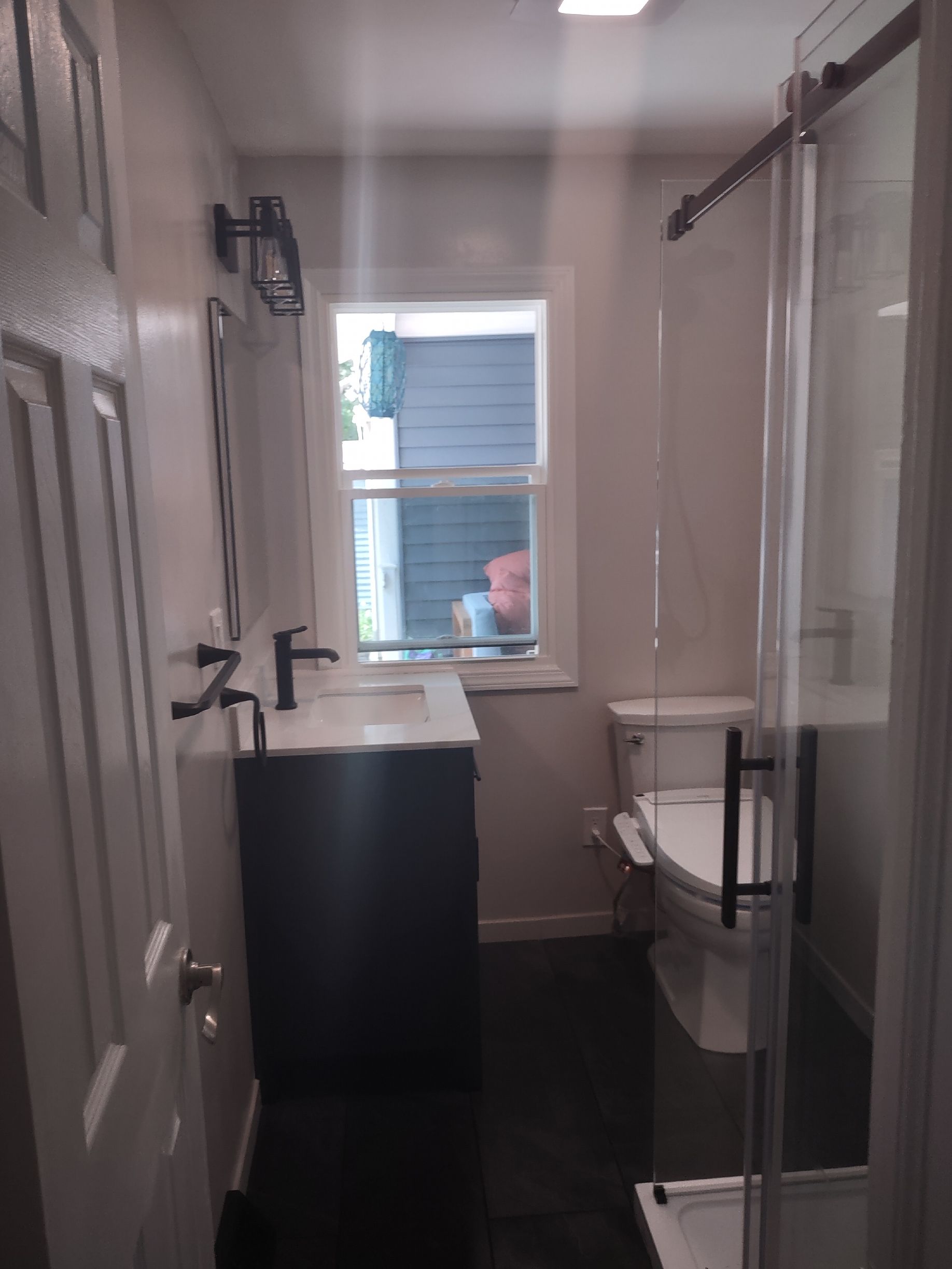 Bathroom with a sink, toilet, shower, and a window. Dark floor and gray walls.