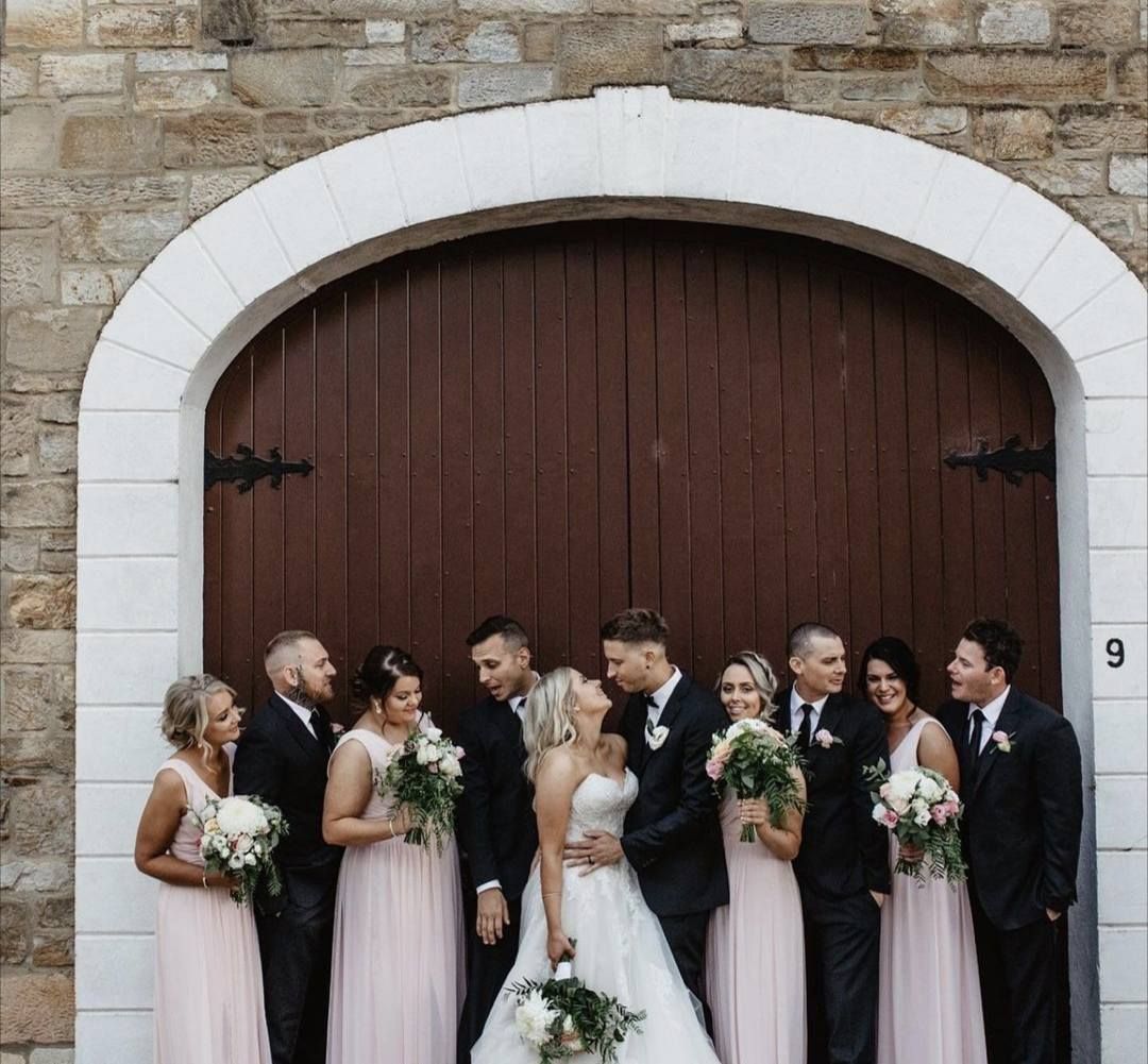 Wedding party posing in front of a wooden door. Bride and groom kiss, bridesmaids in pink, groomsmen in black — Hello Poppy Lane Florist In Albury, NSW