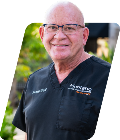 Smiling person in black medical scrubs with glasses, standing outdoors in front of greenery.