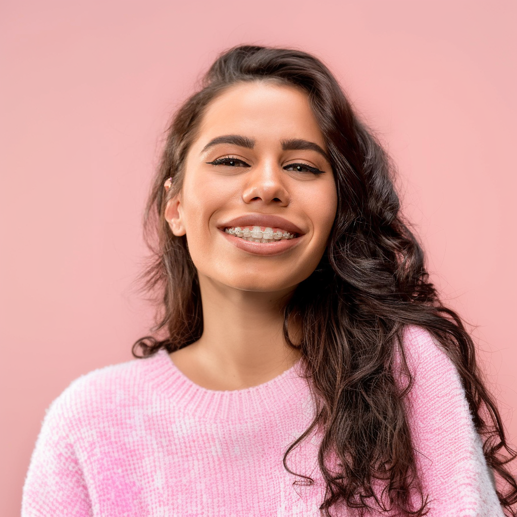 Woman with curly brown hair wearing a pink sweater, smiling, and showing braces against a pink backdrop.
