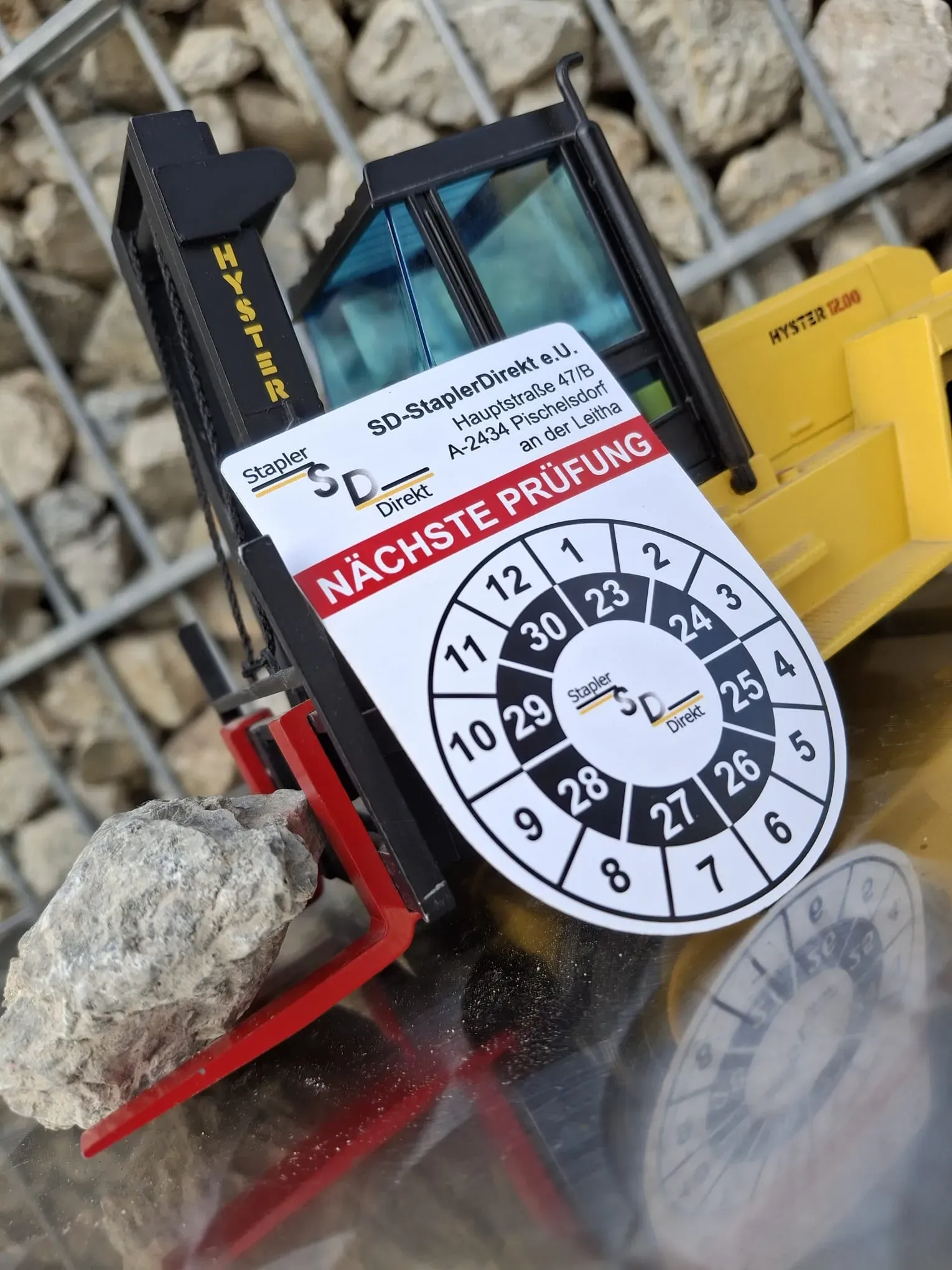 Ein gelb-schwarzer Spielzeuggabelstapler steht vor mit Steinen gefüllten Hintergrund mit Prüfaufkleber „Nächste Prüfung“.