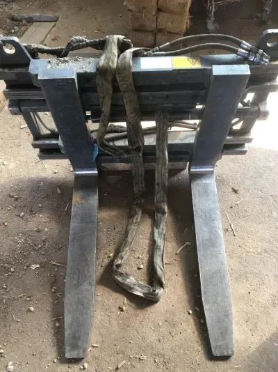 Ein grauer Gabelstapler-Aufsatz mit zwei Gabeln, der auf einem Betonboden ruht und mit einem Stoffgurt überzogen ist.