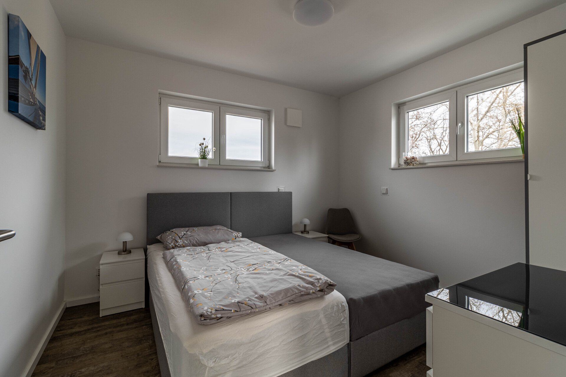 Ein Schlafzimmer mit Bett, Nachttisch, Spiegel und zwei Fenstern.