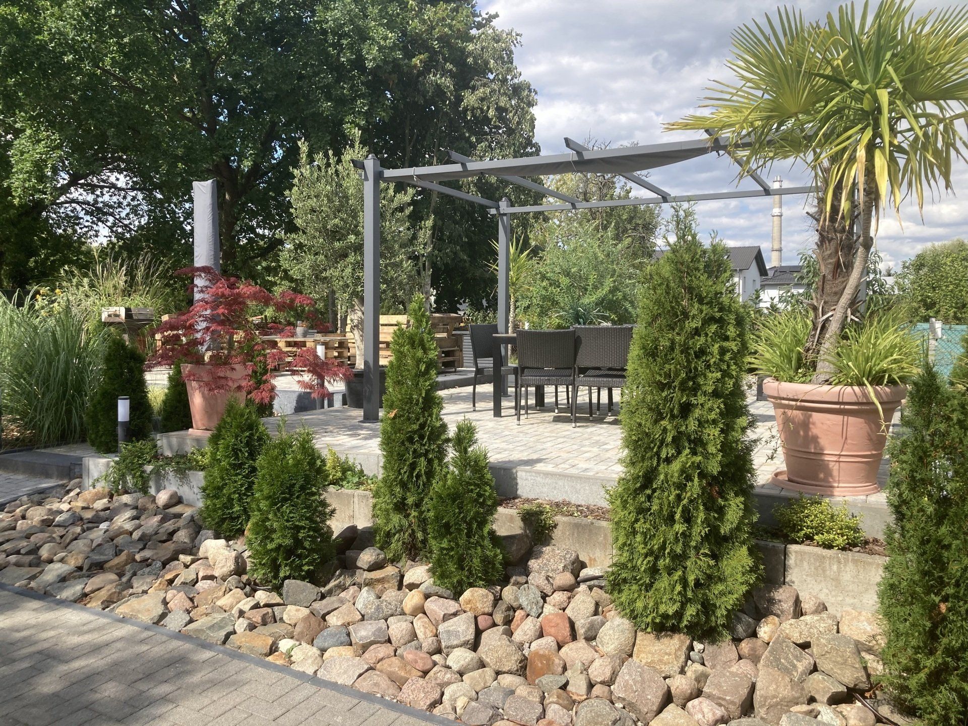 Ein Terrassenbereich mit Pergola, Tischen und Stühlen, umgeben von Bäumen und Felsen.