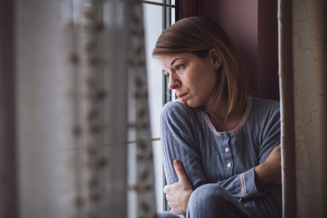 A person in blue loungewear sits by a window, looking out with a contemplative expression.