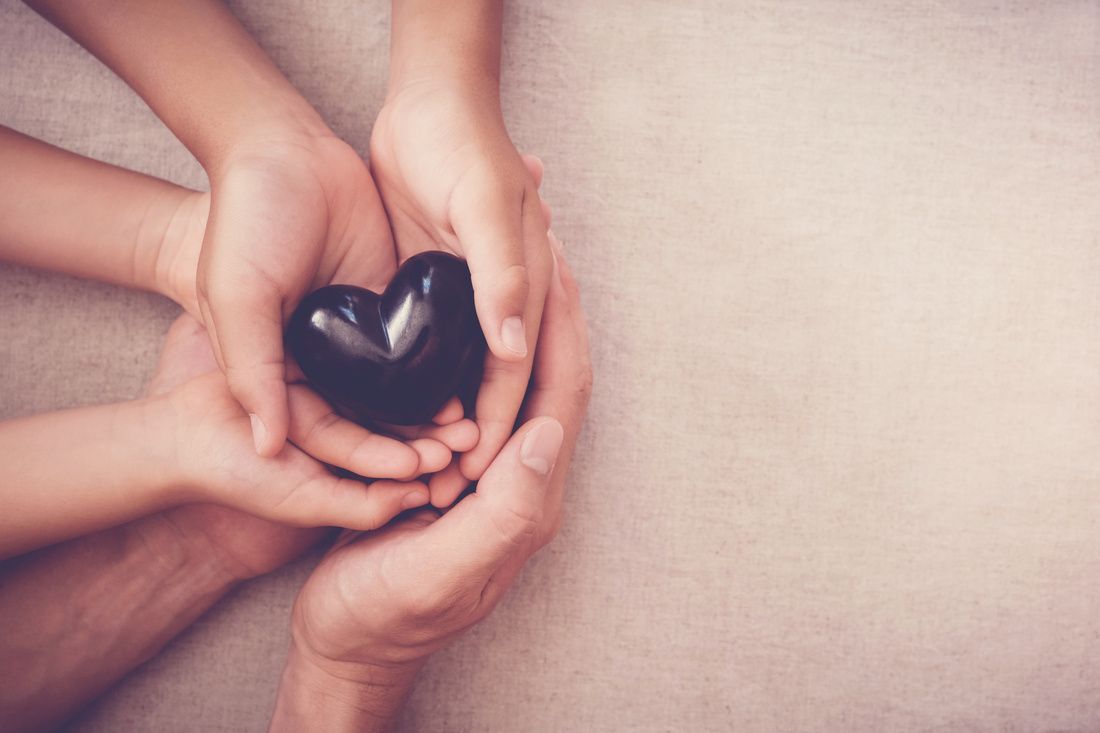 Multiple hands cupping a small, shiny black heart-shaped stone against a neutral, textured background.