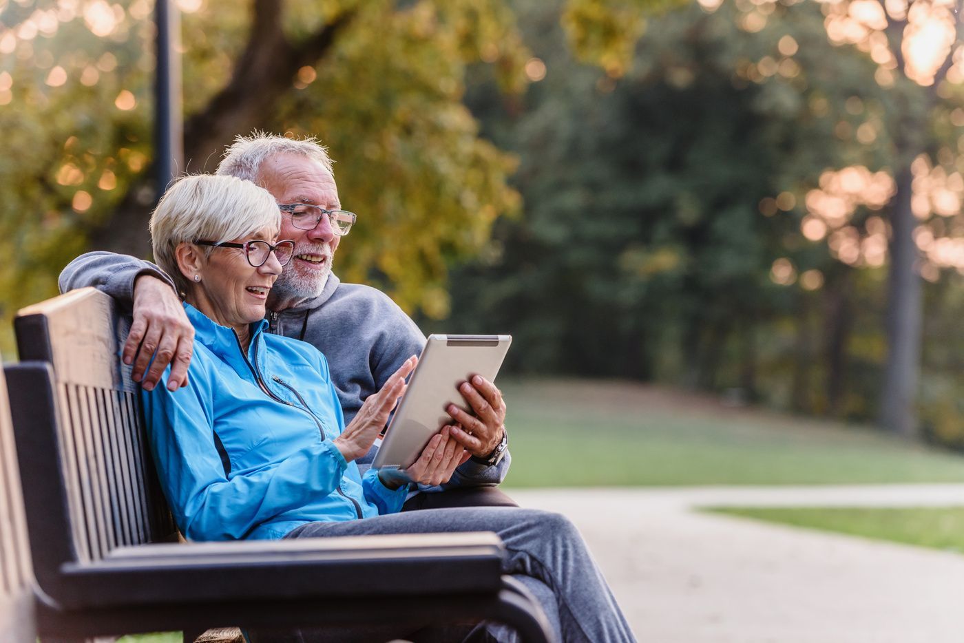Two people sitting on a park bench together, smiling while looking at a tablet held in their hands.