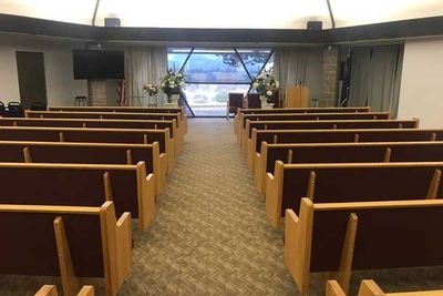 Rows of wooden church pews facing a podium and a large windowed alcove in a room with a tan carpet.