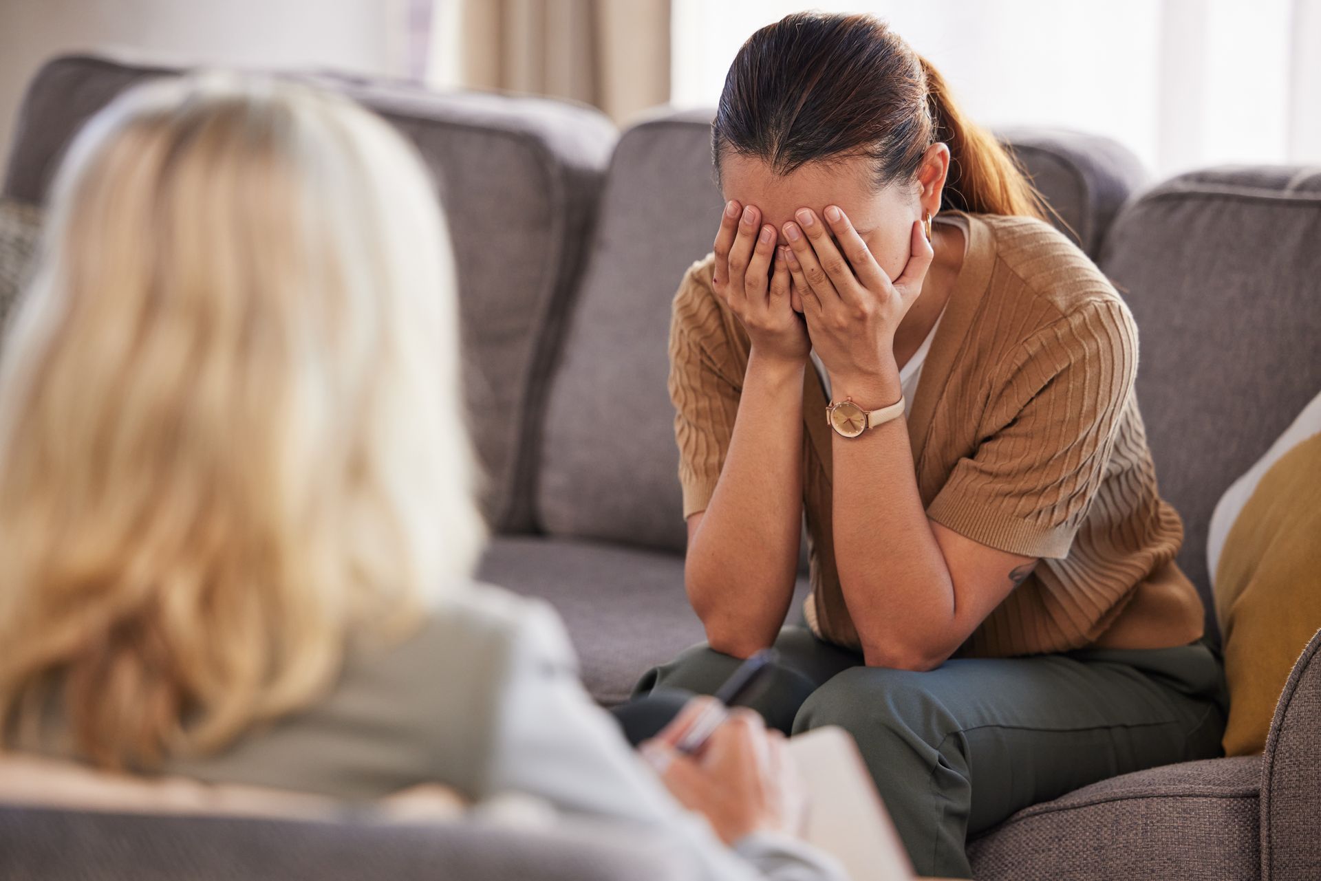 Two women talking on a couch; one covers her face with her hands, appearing upset.