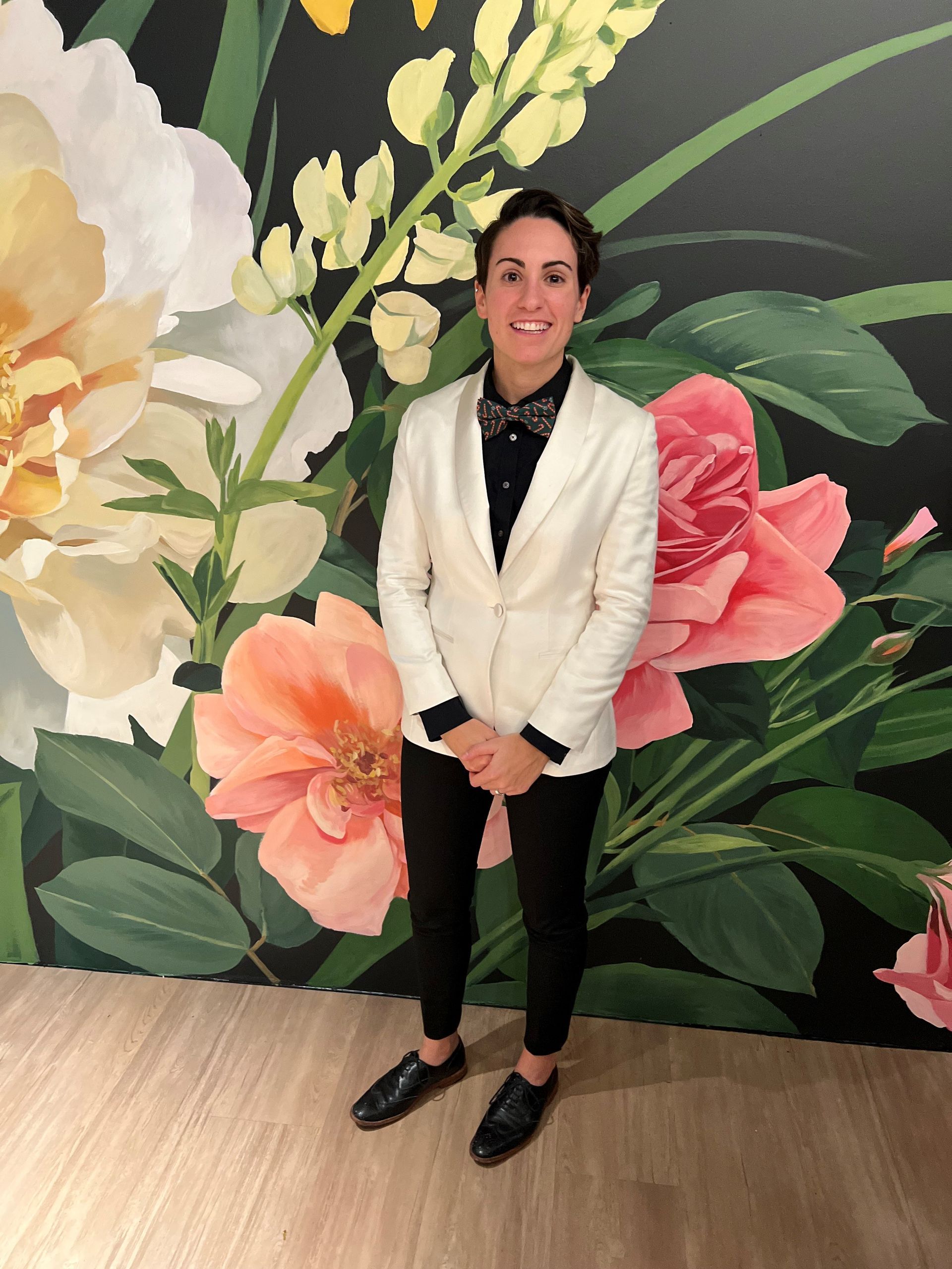 A woman in a white suit is standing in front of a wall of flowers.