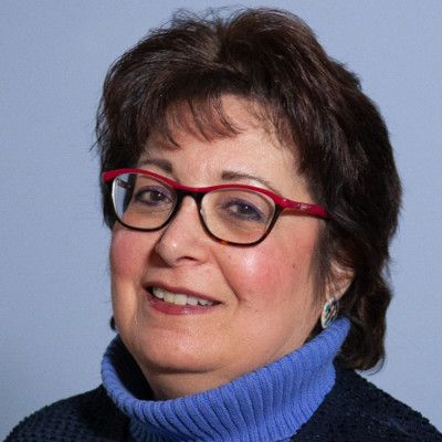 A woman wearing glasses and a blue sweater is smiling.