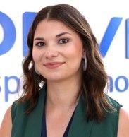 A woman wearing a green vest and earrings is smiling in front of a blue sign.