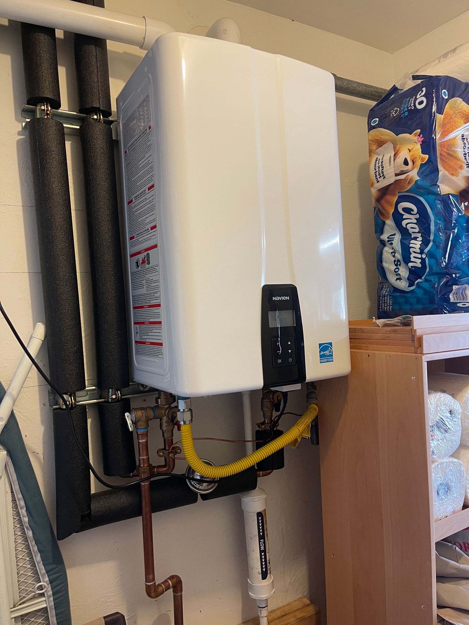 Wall-mounted white water heater with copper piping in a utility closet beside stacked paper towels.