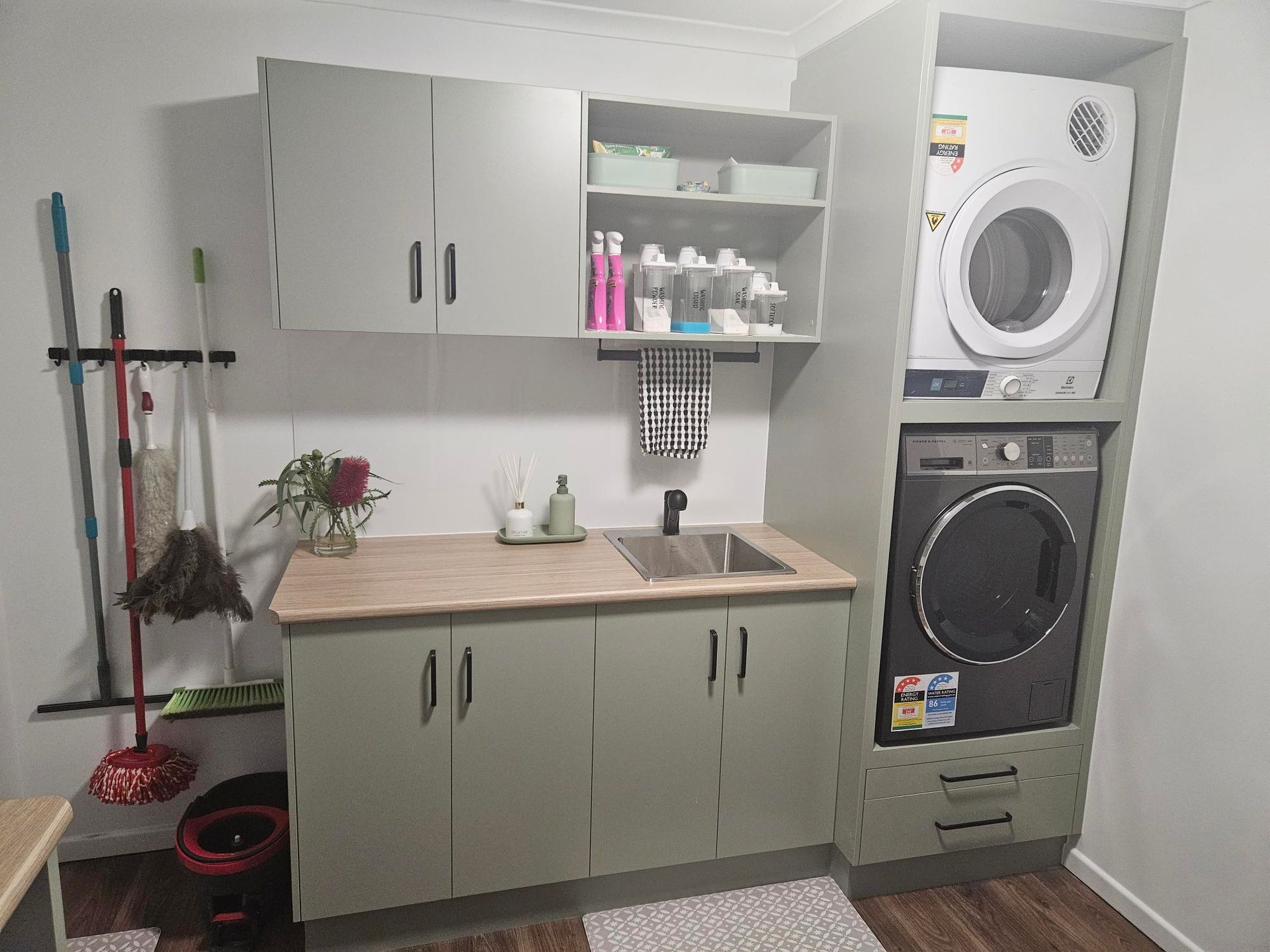 Laundry room with green cabinets, sink, washer, and dryer. Mops and brooms hang on the left.