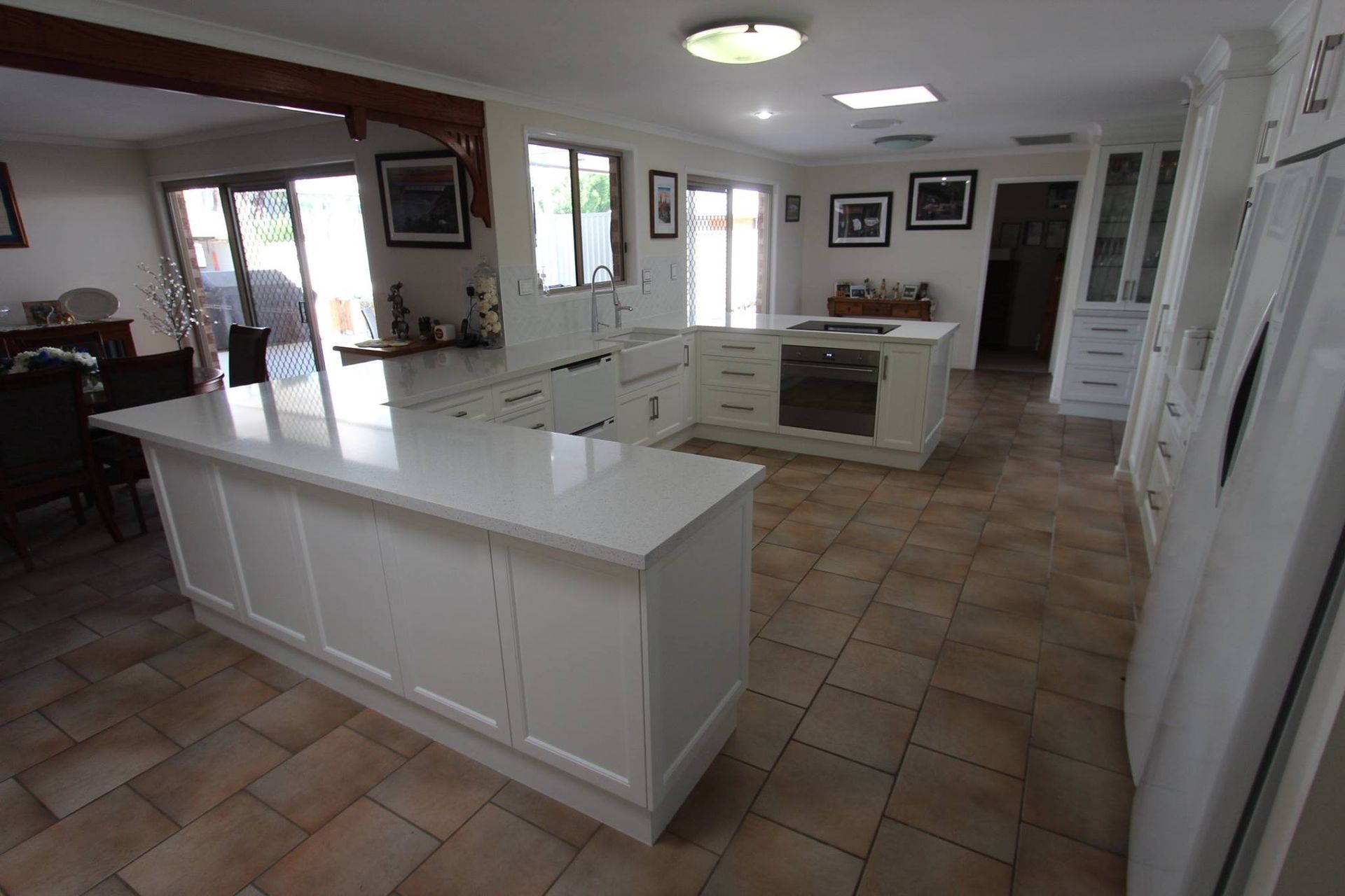 White kitchen with large island and light-colored tile floor. Sliding glass doors and windows provide natural light.