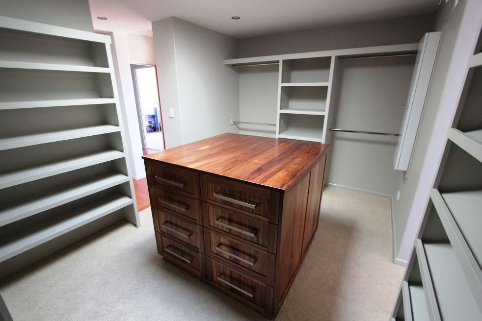 Walk-in closet with shelves and drawers. Wooden island in center. Gray walls and white shelves.