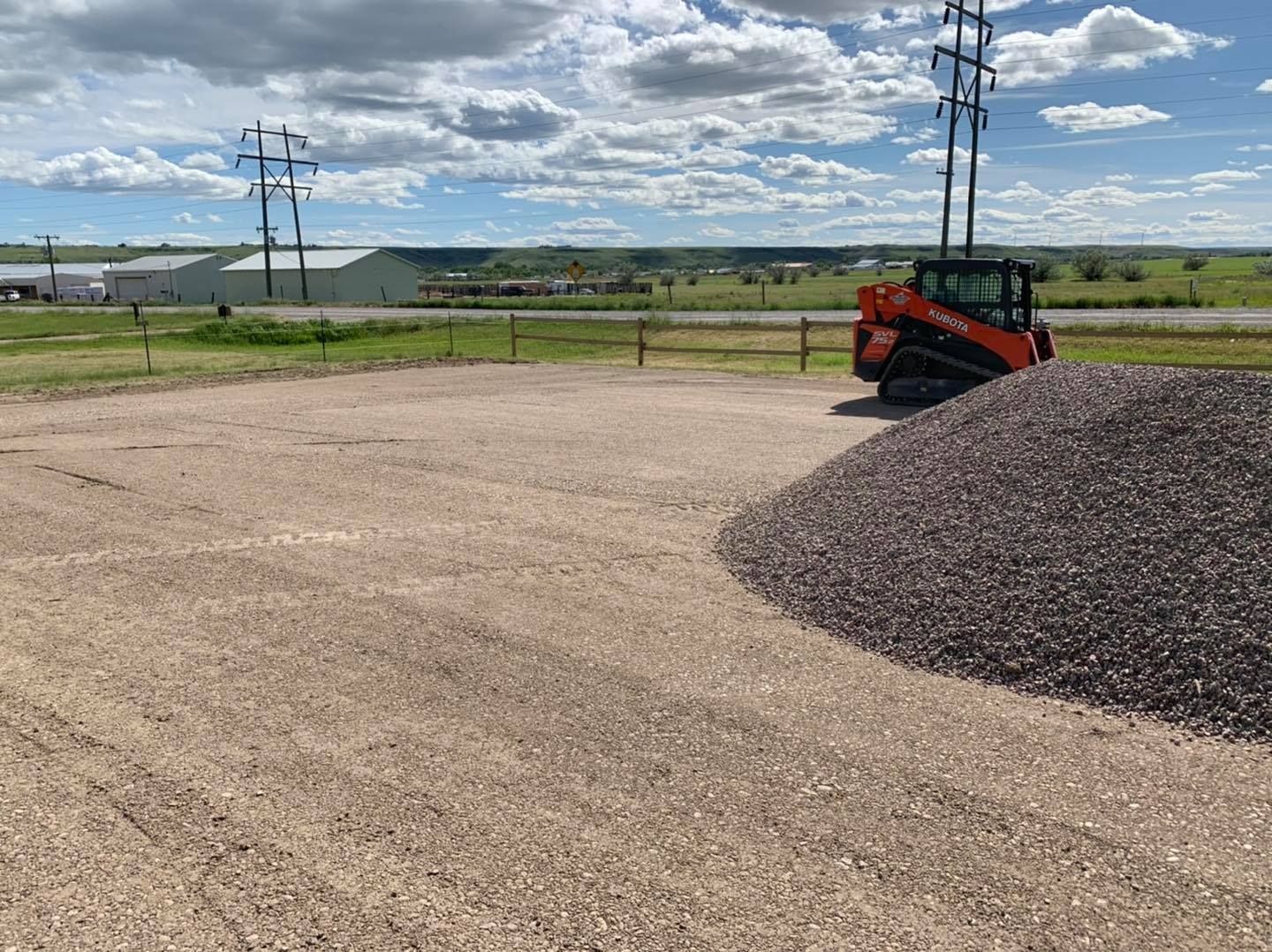 An orange skid-steer loader sits next to a large pile of gravel on a flat, gravel-covered lot under a cloudy sky.