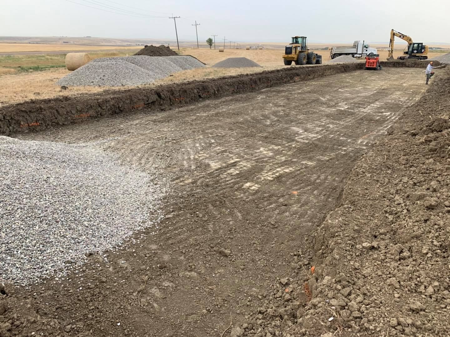 Construction equipment at a site with piles of gravel and a prepared dirt foundation in an open landscape.