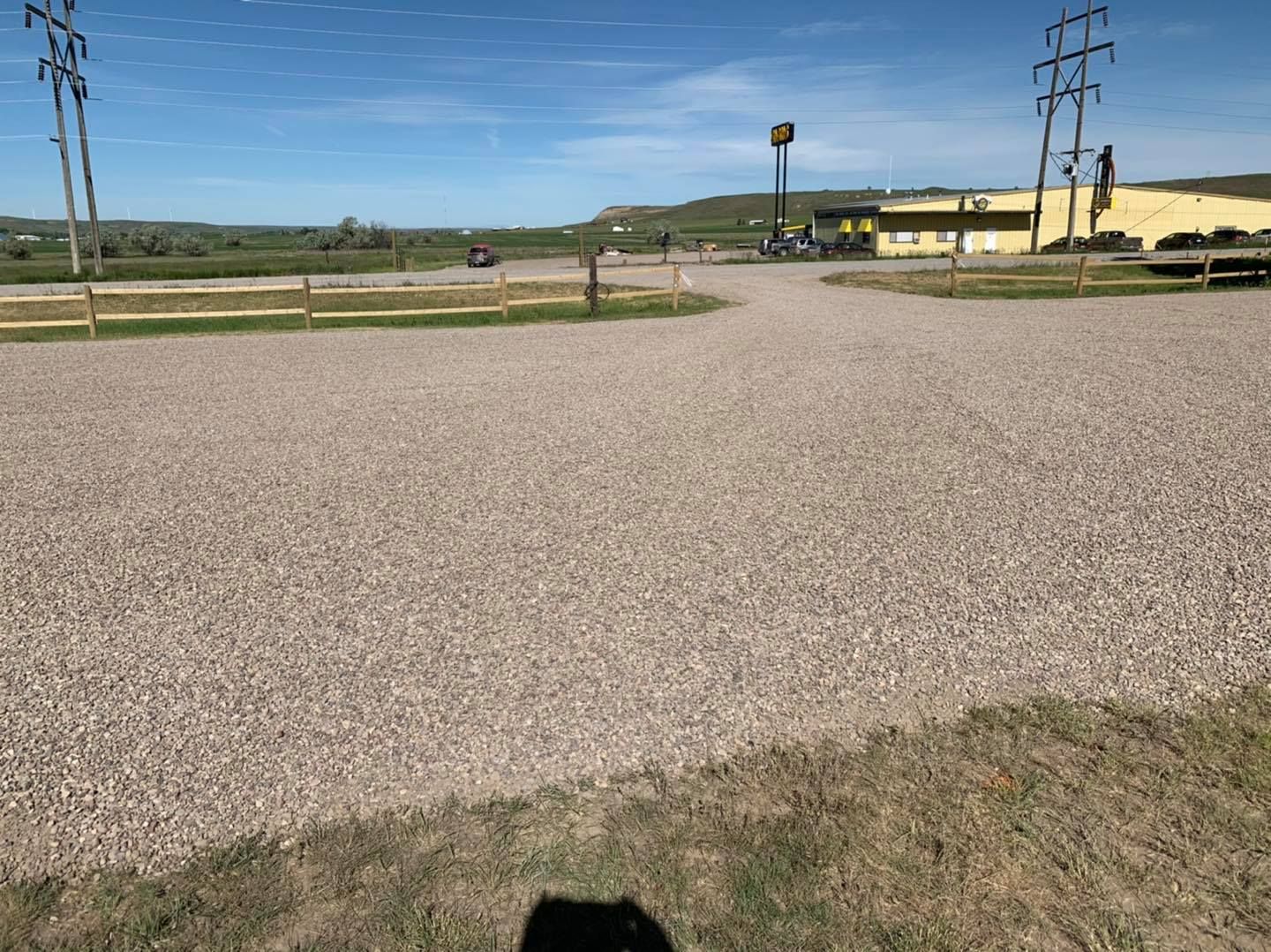 A wide, gravel parking lot under a clear blue sky, with utility poles and a yellow building in the distance.