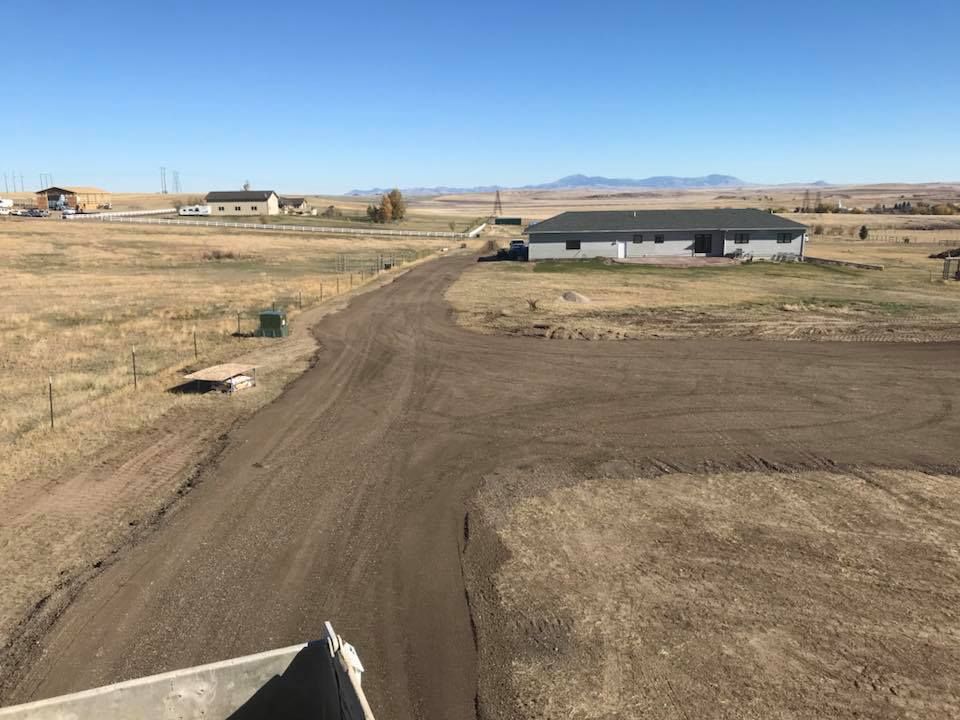 An unpaved dirt driveway curves through a dry, grassy field toward a house under a clear blue sky.