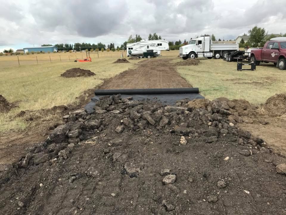 A wide dirt path is under construction in a grassy field, with a layer of black landscape fabric being unrolled.