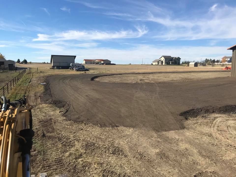  A freshly graded dirt driveway curves through an open, dry field under, with rural buildings in the distance.