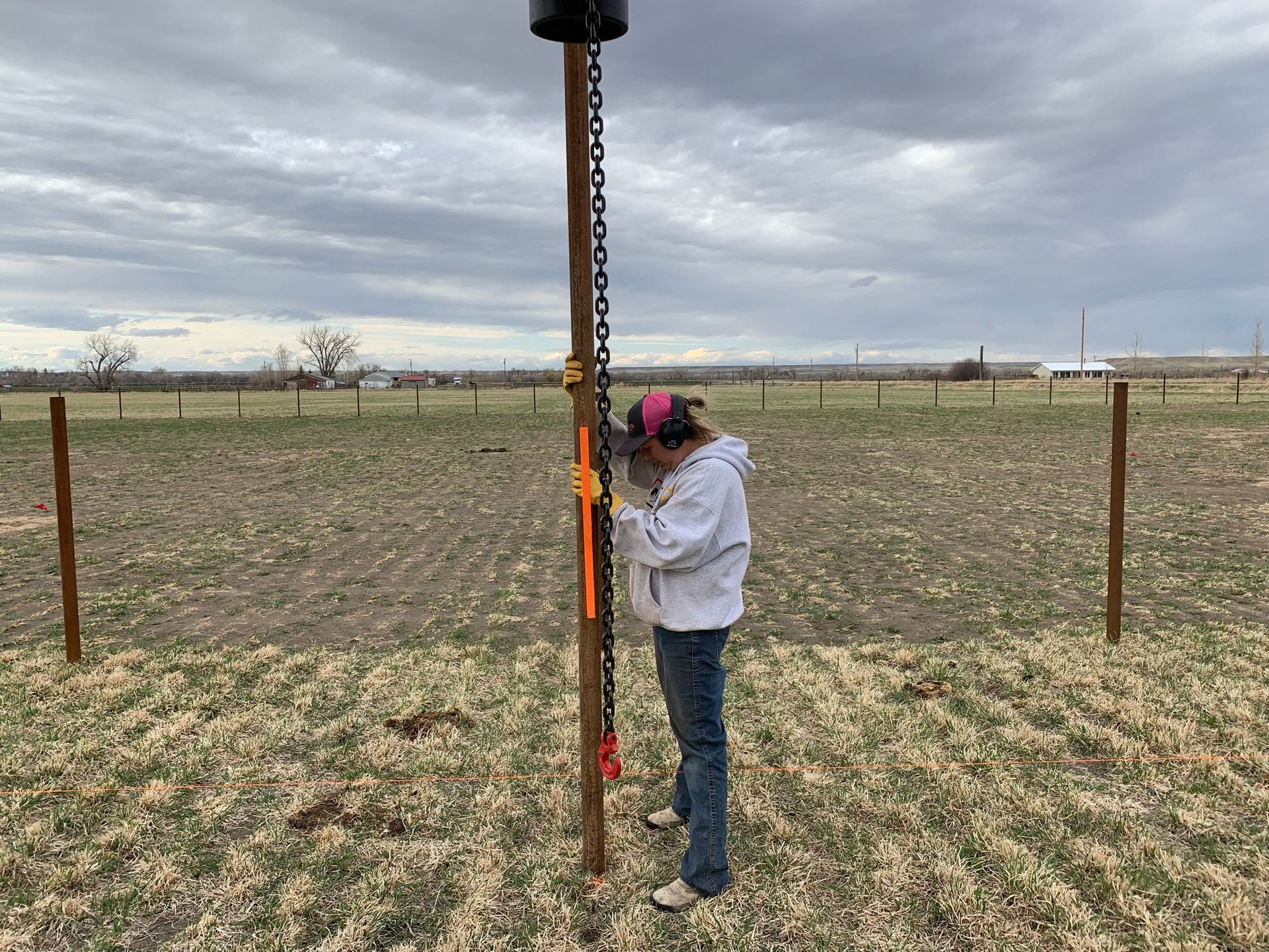 A person in a gray hoodie and blue jeans stands in a field, inspecting a wooden fence post with a metal chain attached.