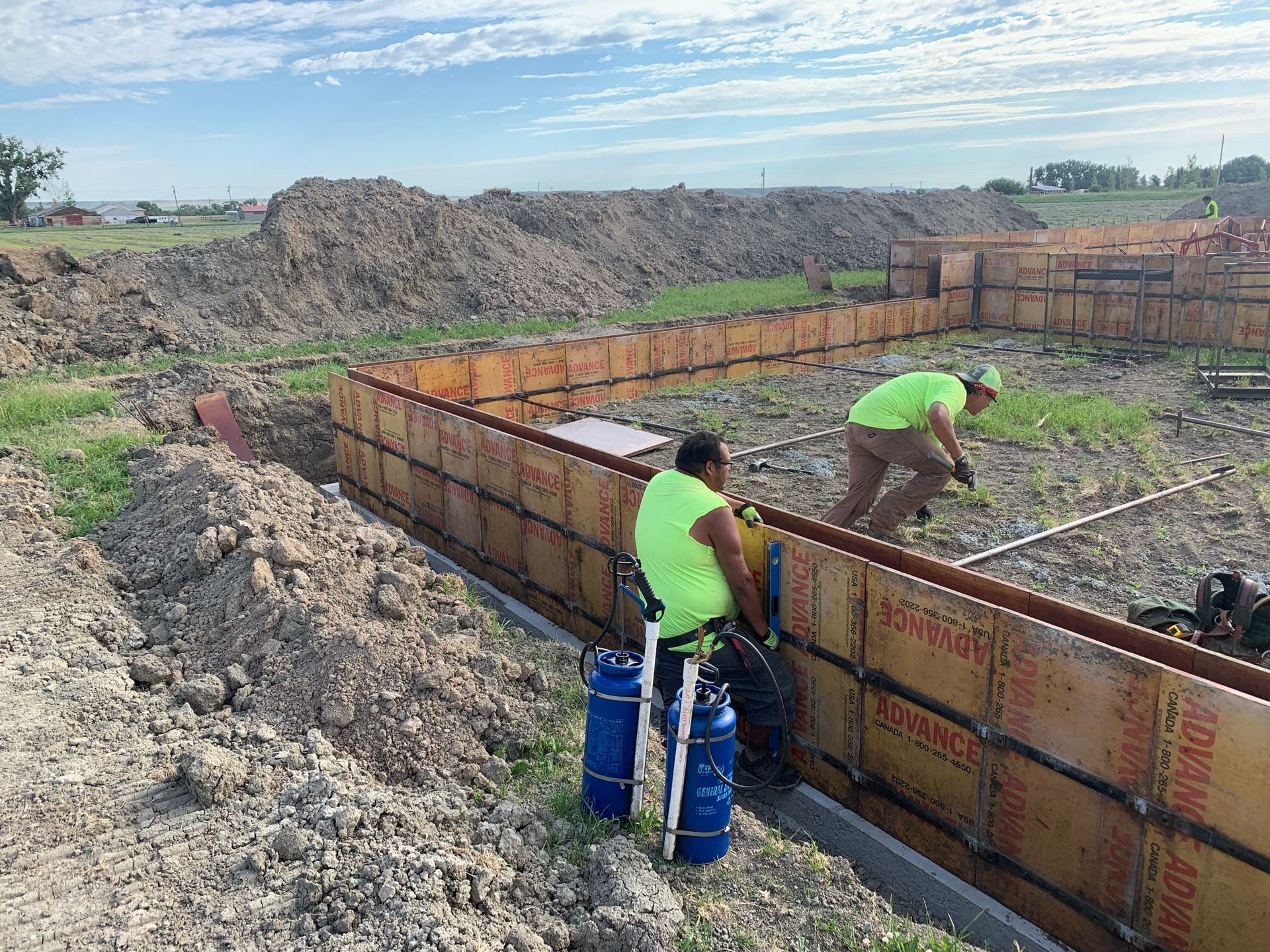 Two construction workers in neon shirts assemble wooden concrete forms at a dirt construction site.