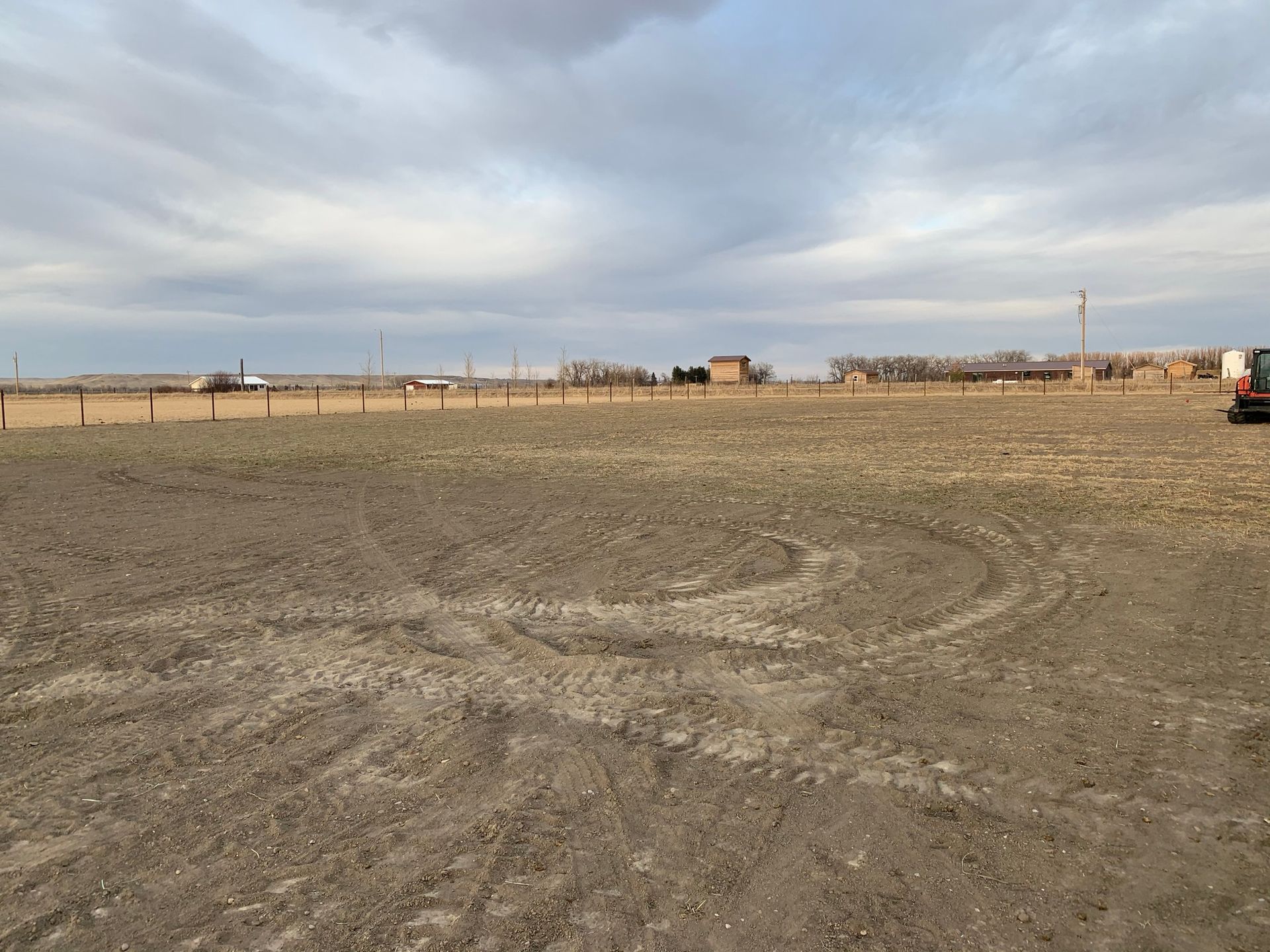 A wide, flat, dirt field under a cloudy sky, with a fence line and distant farm structures in the background.