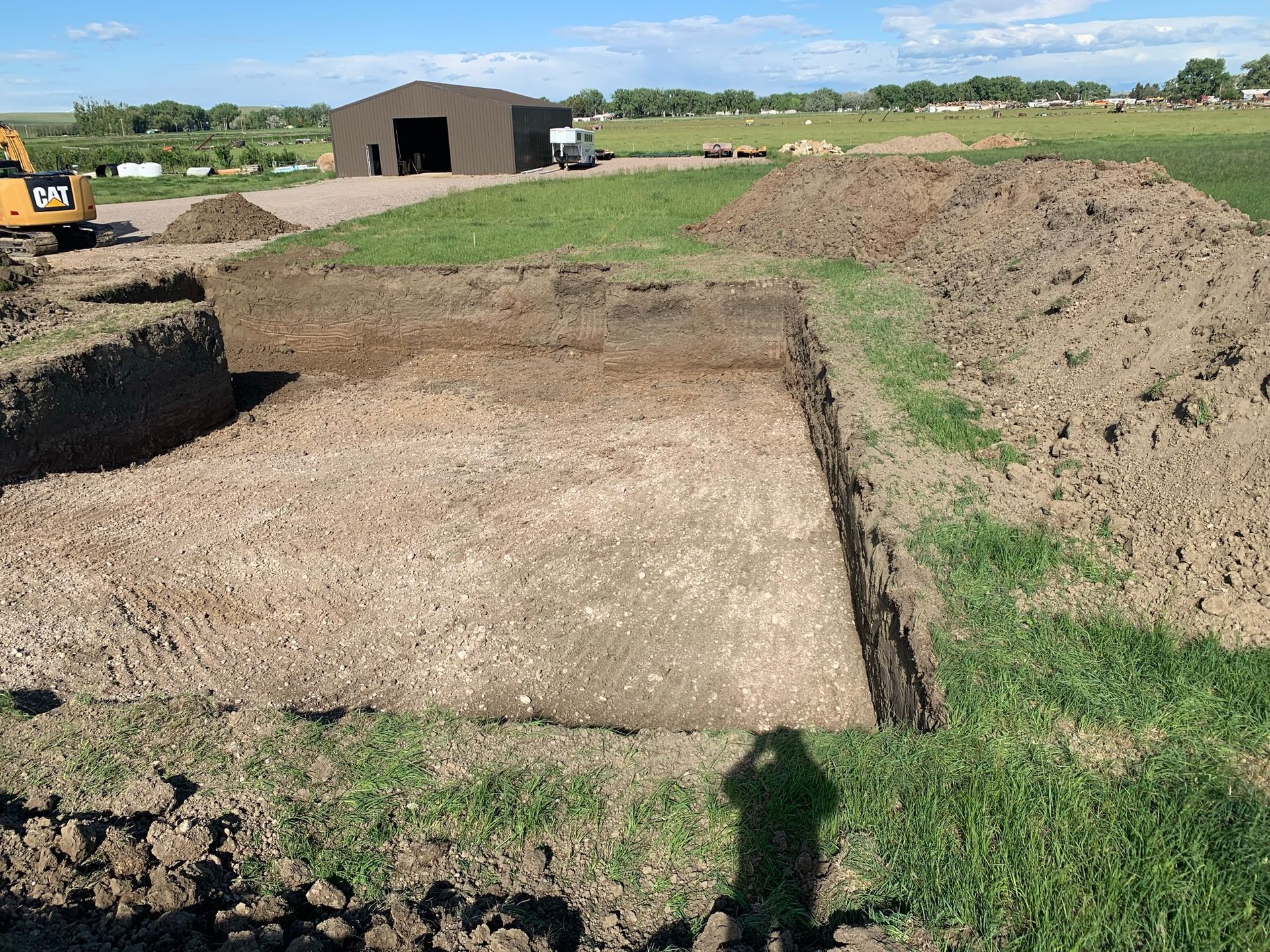 A large, rectangular construction excavation pit in a grassy field with piles of dirt and a shed in the background.