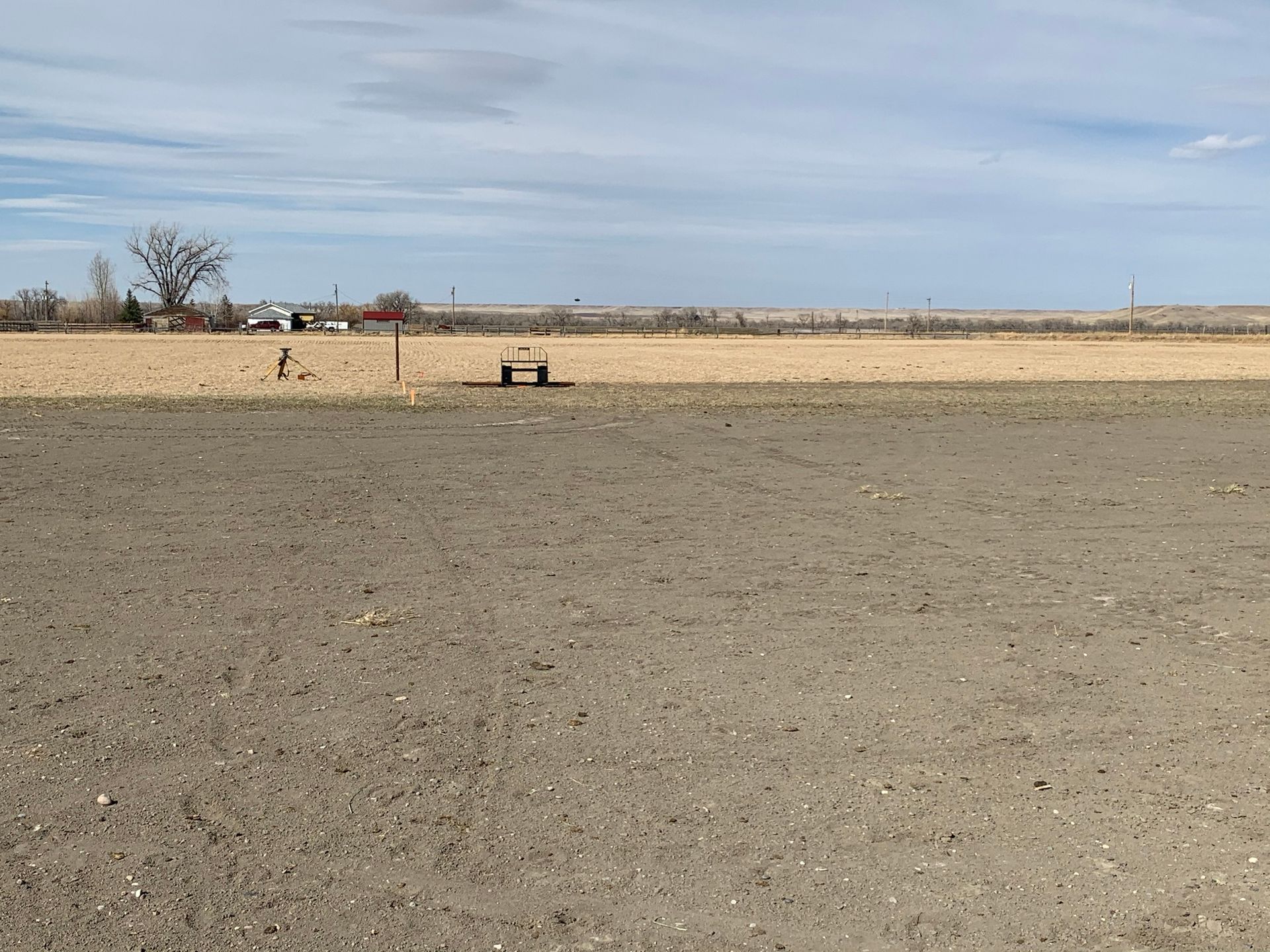 A flat, barren agricultural field under a cloudy sky with a small structure and bare trees in the distance.
