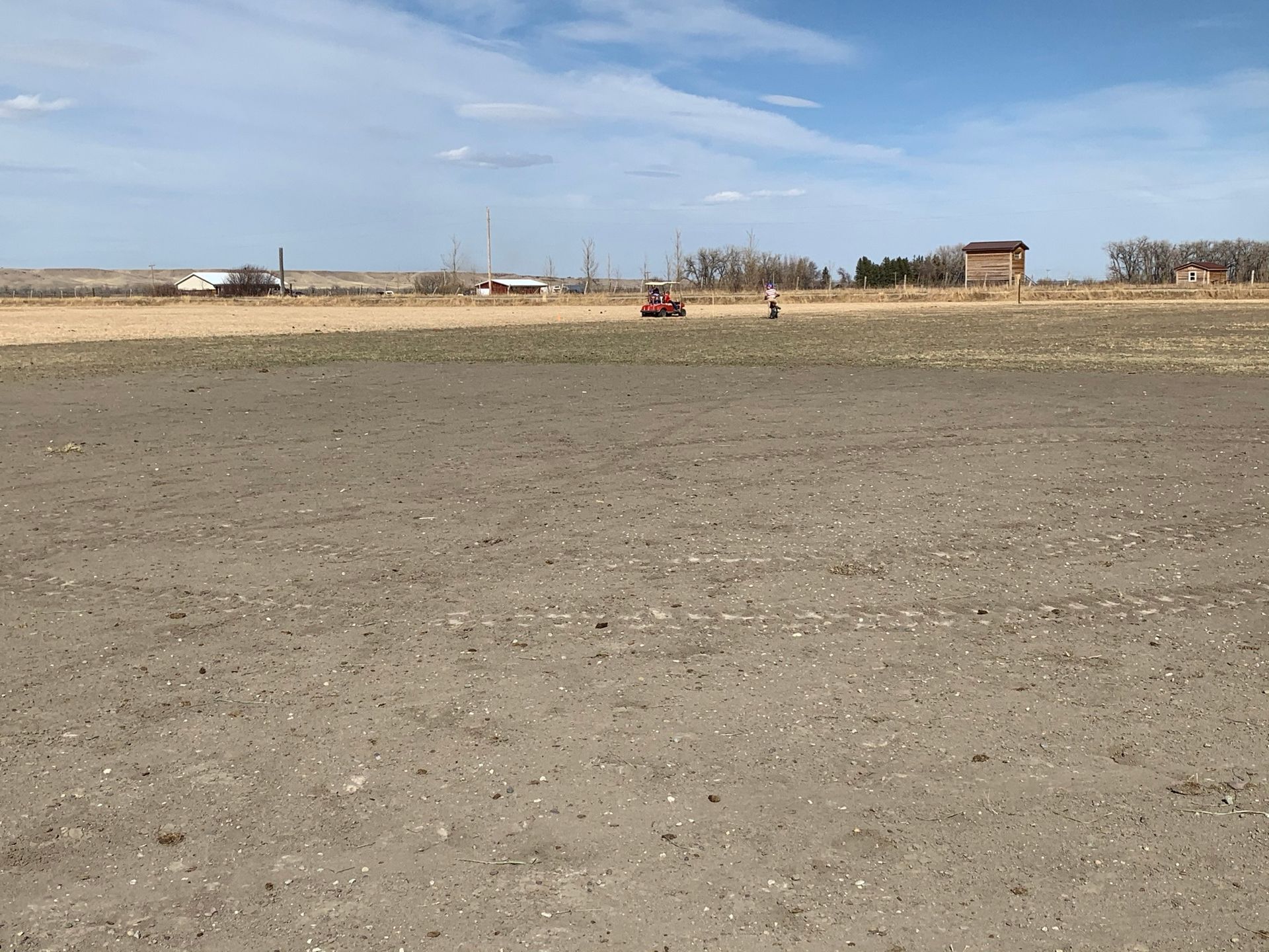 A person in a red vehicle drives across a flat, open, tilled field under a cloudy, bright sky.