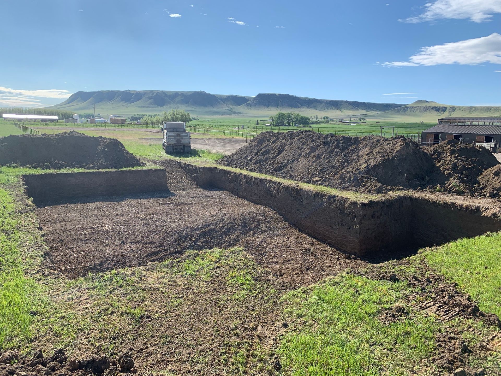 A deep, rectangular excavation in a grassy field with piles of excavated dirt nearby, under a bright, mountainous horizon.